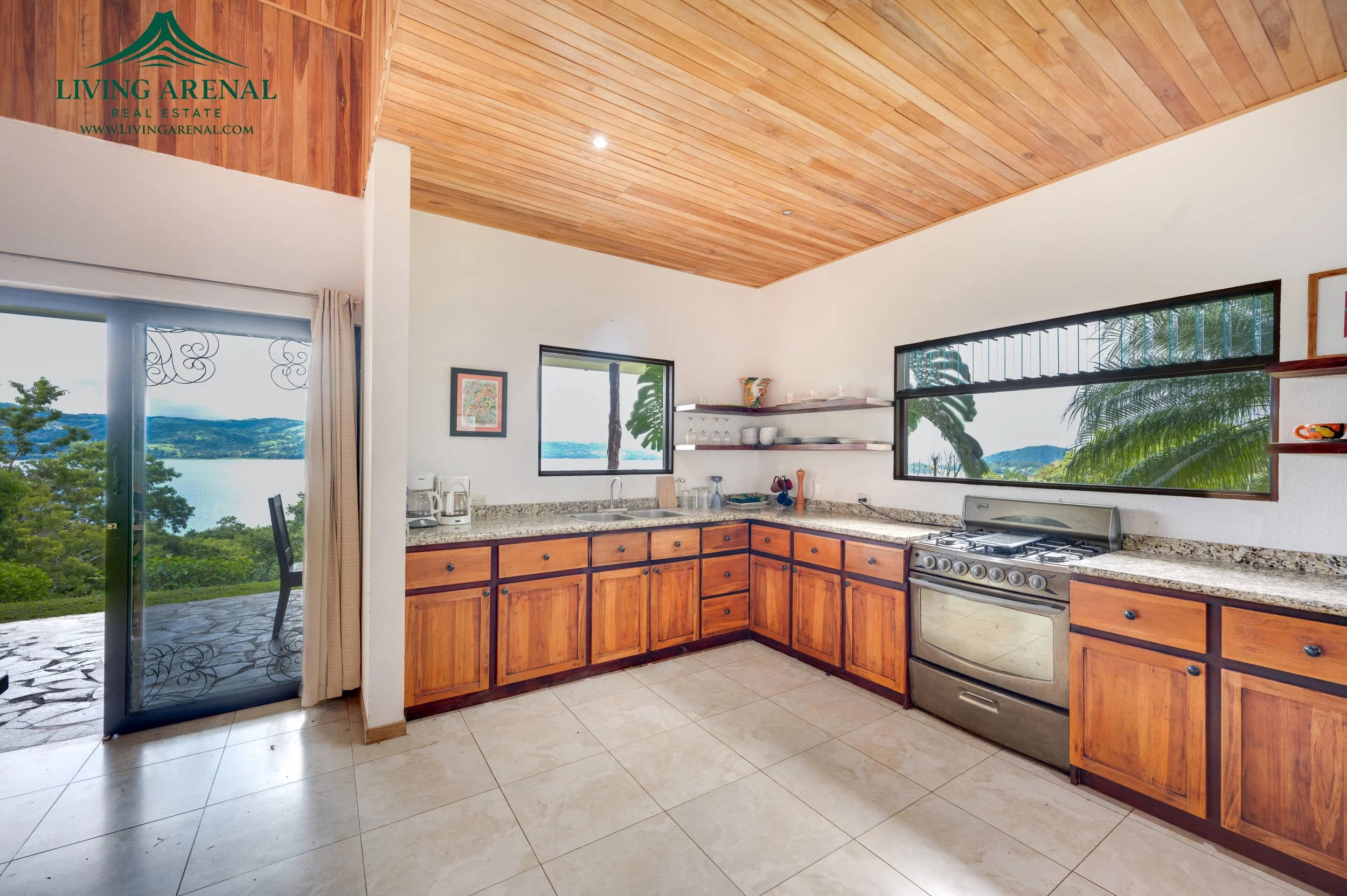 Kitchen with wooden cabinets, granite countertops, stainless steel stove, open shelves with dishes, large windows overlooking a lake and mountains.