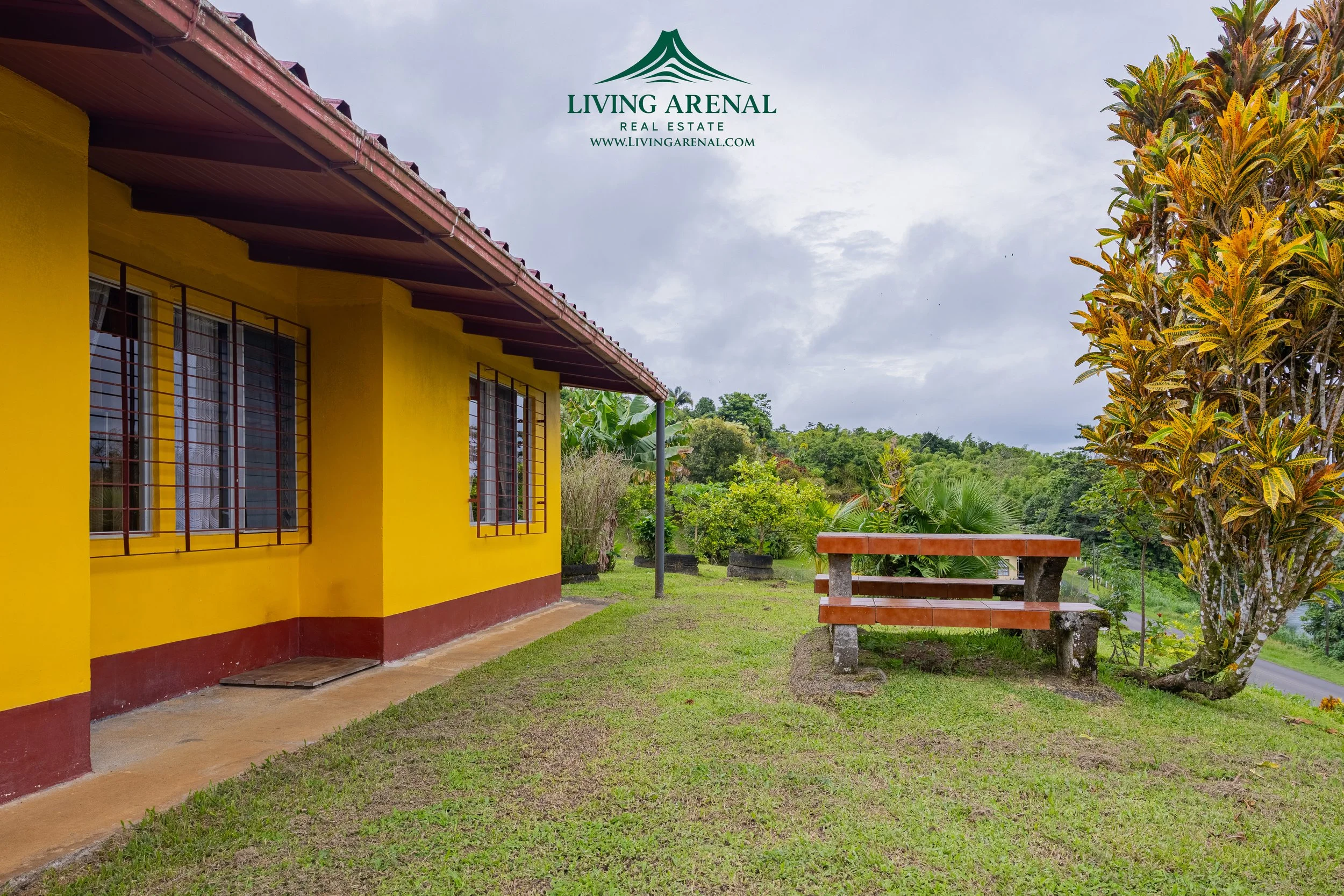 A yellow house with brown trim and a red and brown bench outside, surrounded by green grass, trees, and bushes, with cloudy sky overhead.