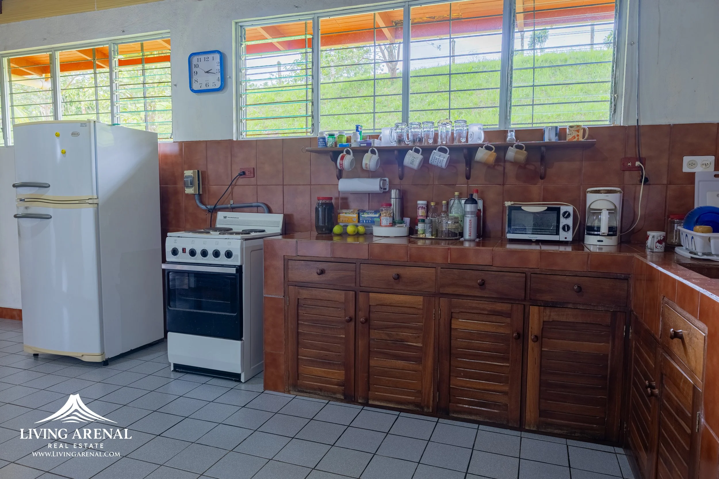 Kitchen with brown tiled walls, wooden cabinetry, a white refrigerator, a small oven, and various small appliances on the counter including a toaster oven and a coffee maker. There are open windows with a view of greenery outside, and shelves with ha