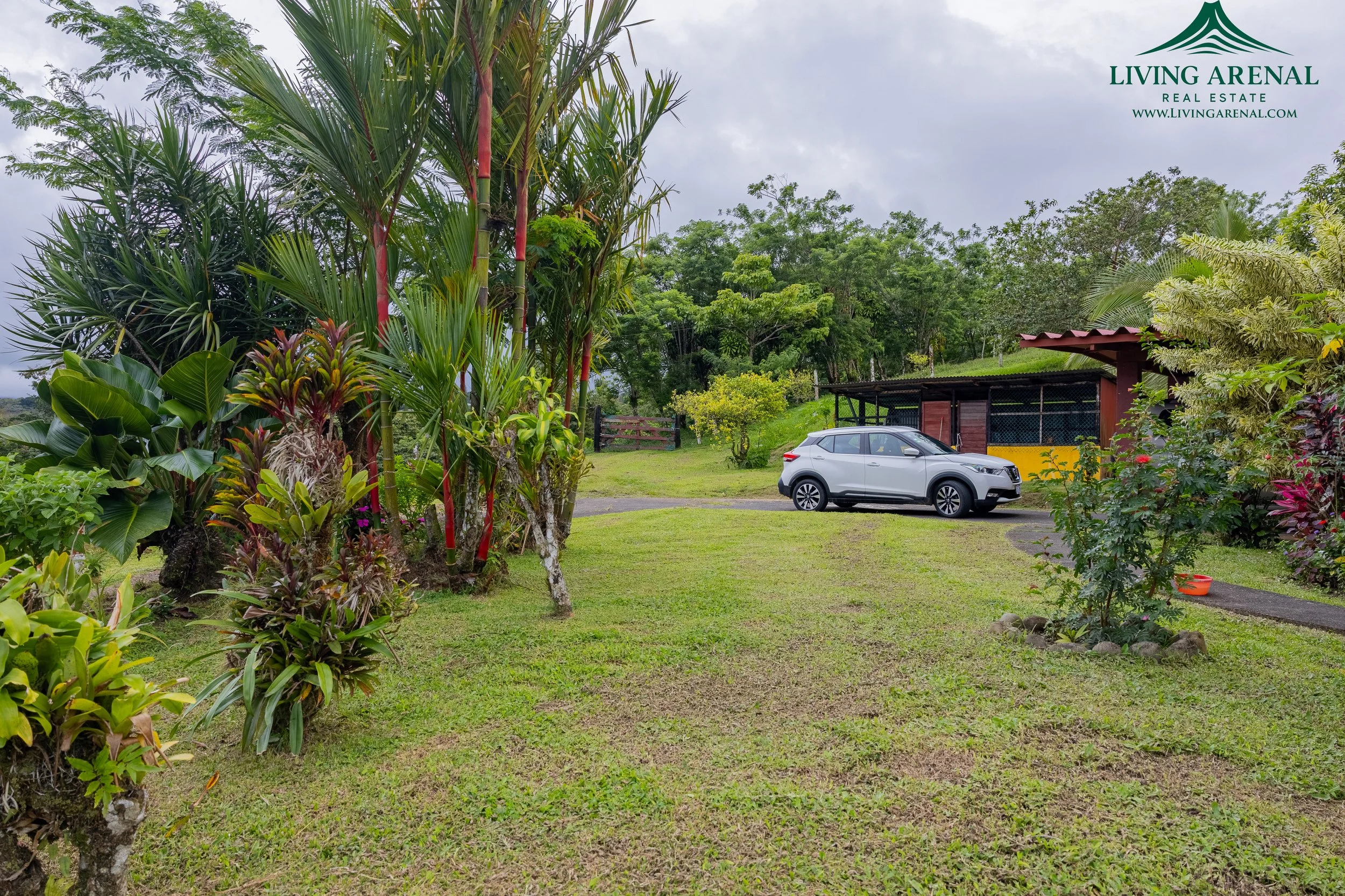A lush, green yard with various tropical plants and trees. A white SUV is parked near a rustic wooden structure with a red roof. The sky is overcast, and there is a small dirt path leading to a gate in the background. The image has a logo in the uppe