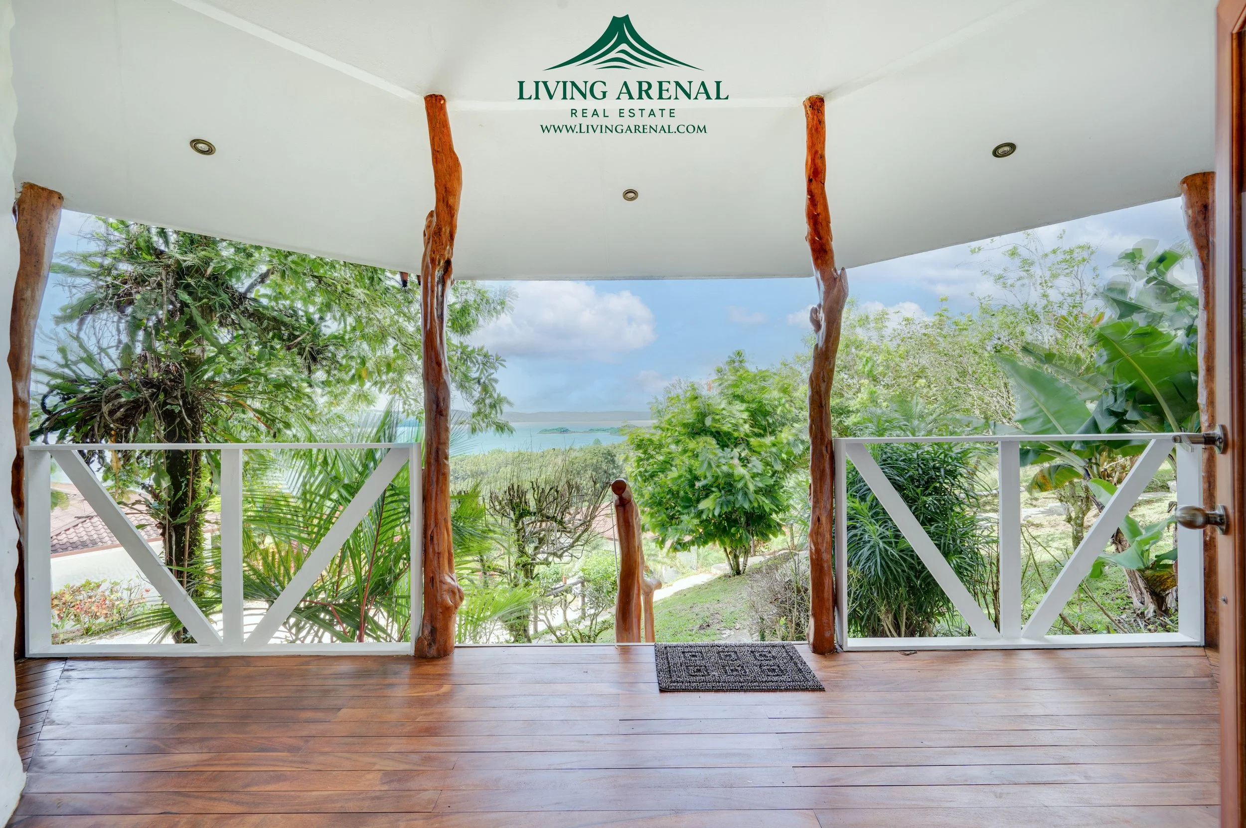 View from a porch with wooden floors, white railing, and rustic tree trunks supporting the ceiling, overlooking a lush tropical landscape with trees and shrubs, and a body of water in the distance.