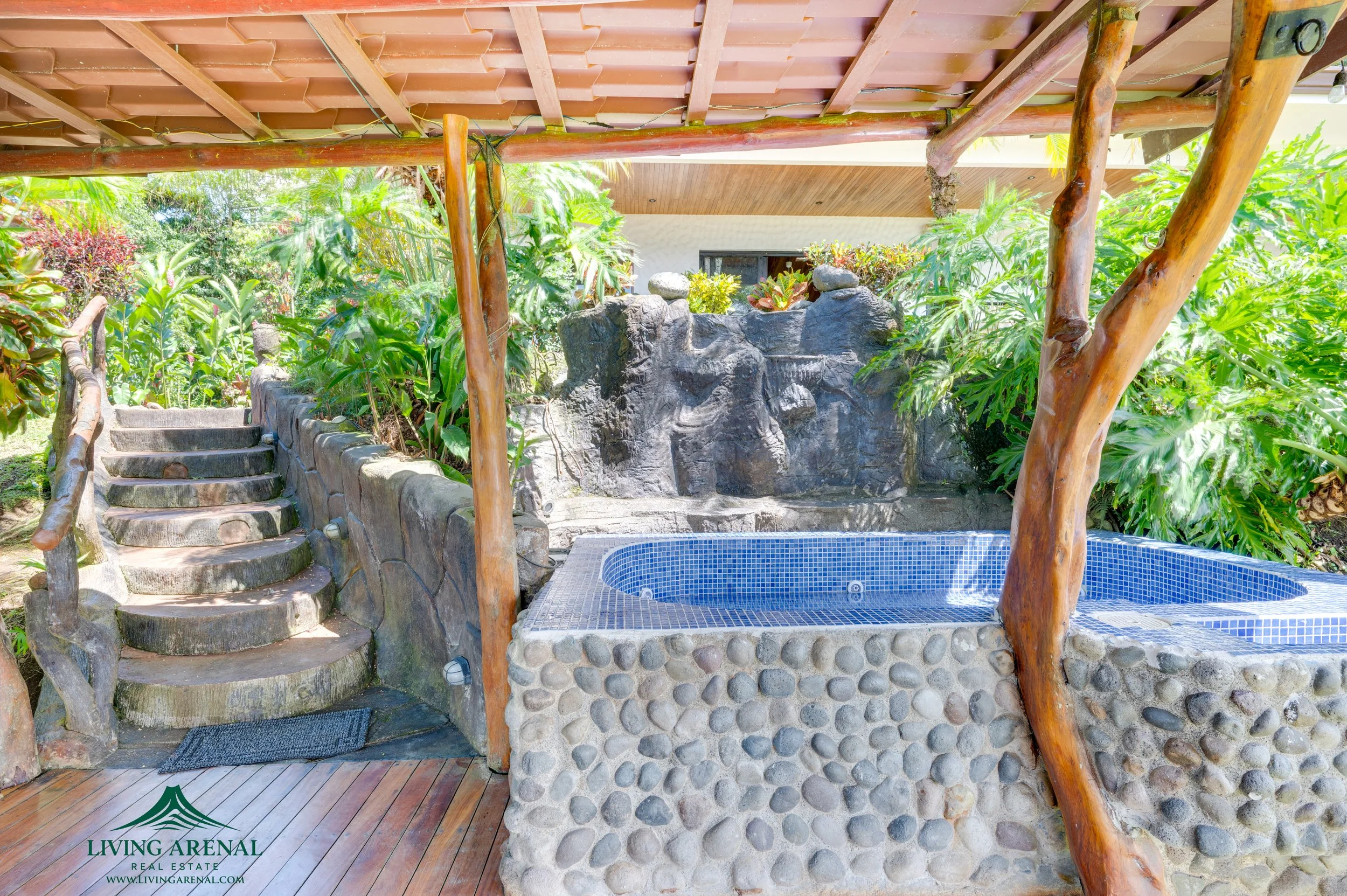 Outdoor hot tub with a pebble stone exterior, set in a lush, tropical garden with green plants and rocks, covered by a wooden roof with wooden support beams.
