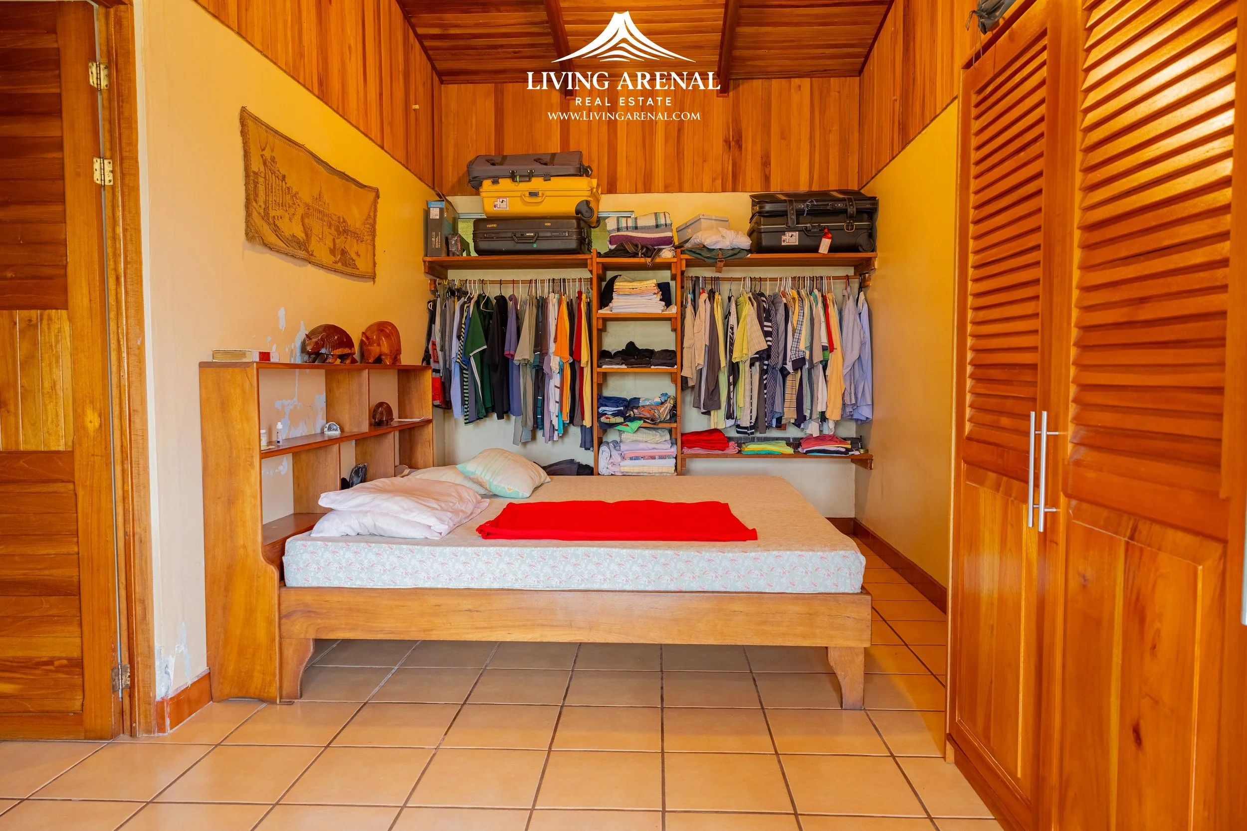 A bedroom with wooden walls and a bed with pillows and a red blanket. An open closet with hanging clothes, folded clothes, suitcases, and bags on top. A wooden wardrobe is on the right side. There is a decorative wall hanging above the bed and a wood