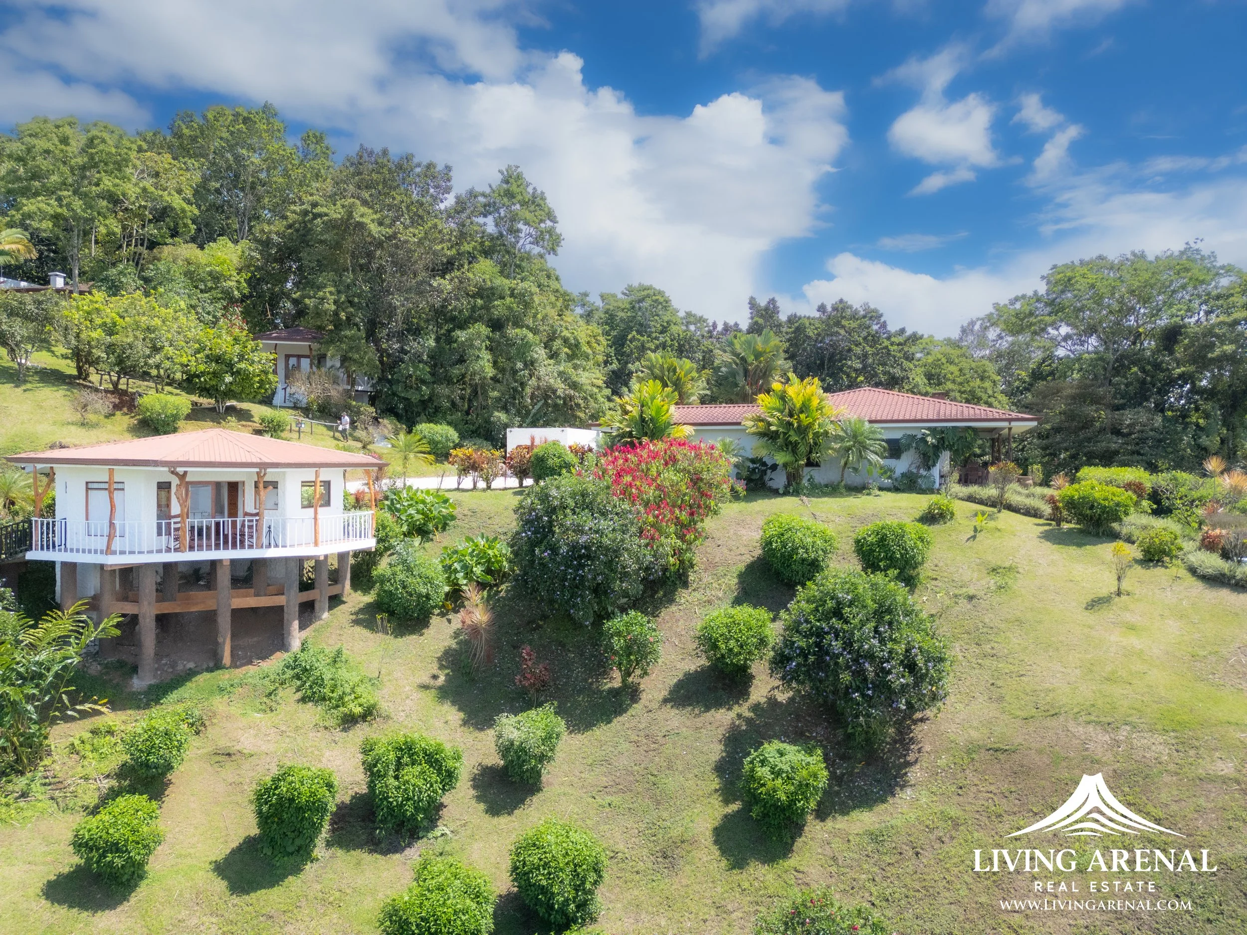 A hillside landscape with two white houses and lush green trees and bushes under a partly cloudy blue sky.