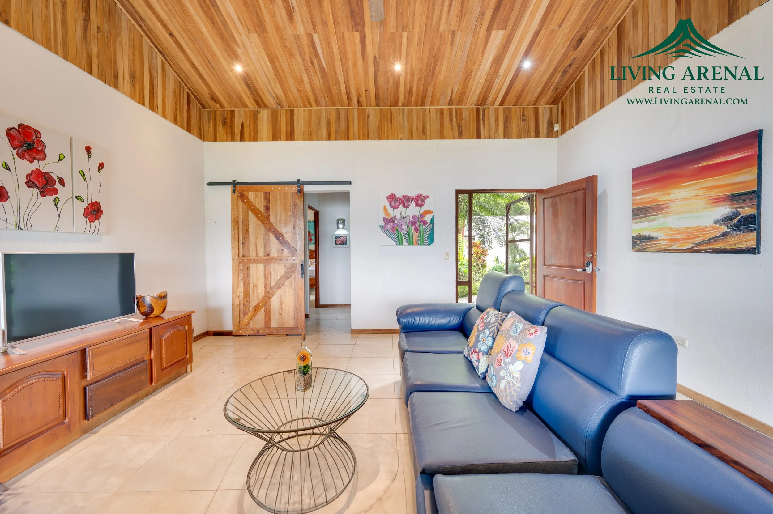Living room with a blue sofa, wooden ceiling, artwork on white walls, a sliding barn door, TV on wooden console, glass-topped coffee table, open door leading to a garden.