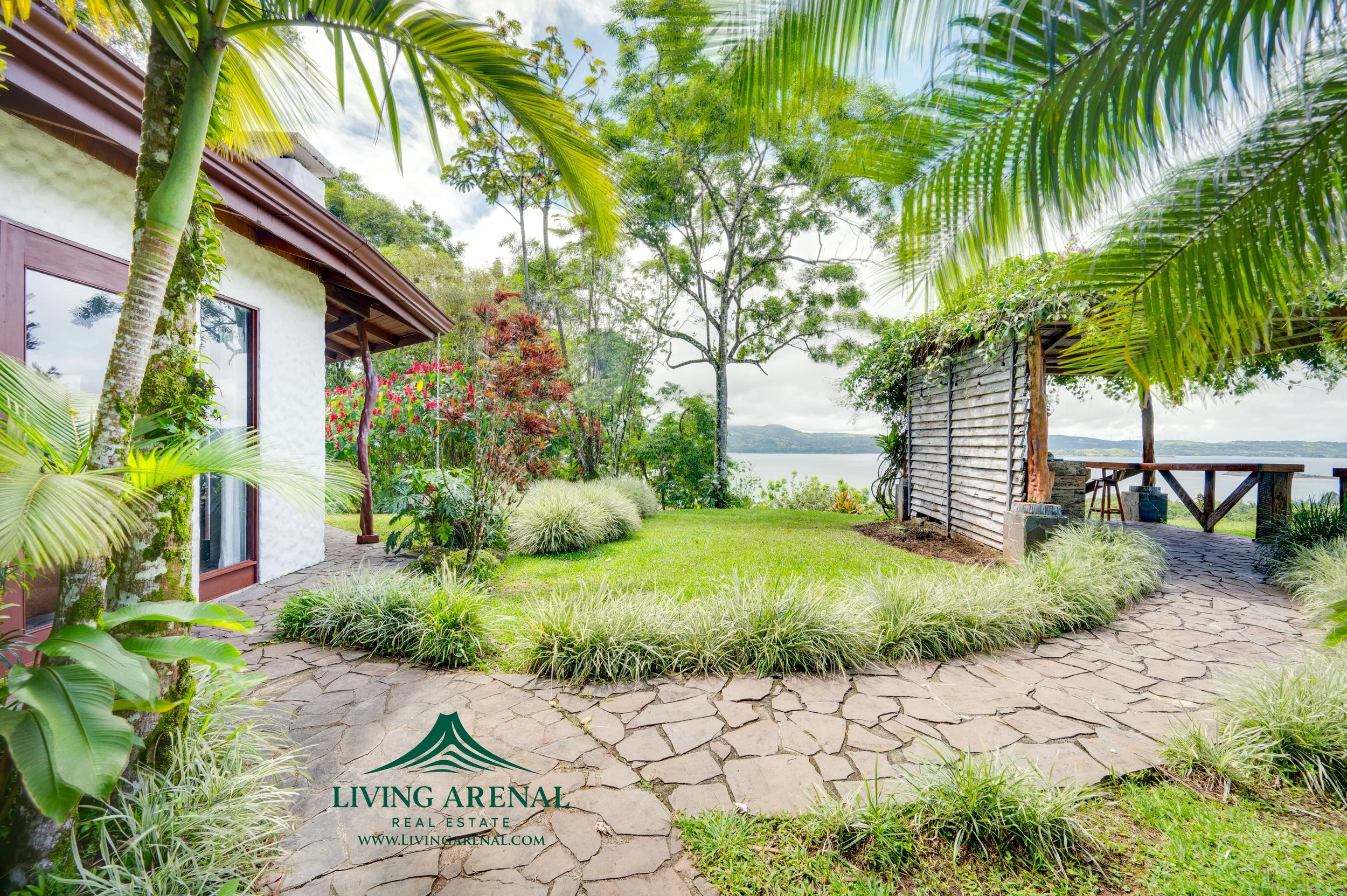 A lush green garden with a stone pathway, tropical plants, trees, and a view of the water in the background. There is a small white house on the left and a wooden structure on the right, both surrounded by greenery.