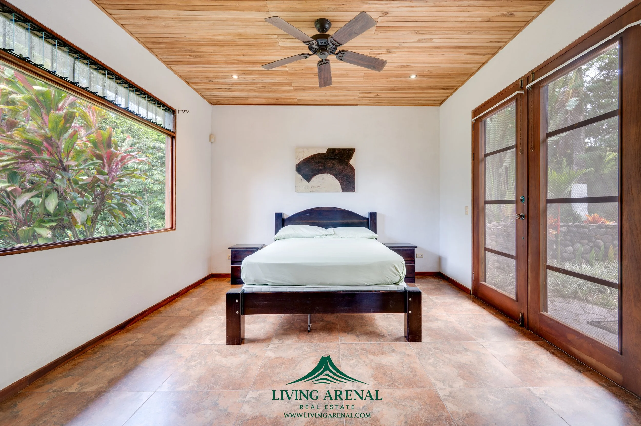 A bedroom with a wooden ceiling, large windows, a wooden door, a ceiling fan, a bed with light-colored linens, and abstract artwork on the wall.