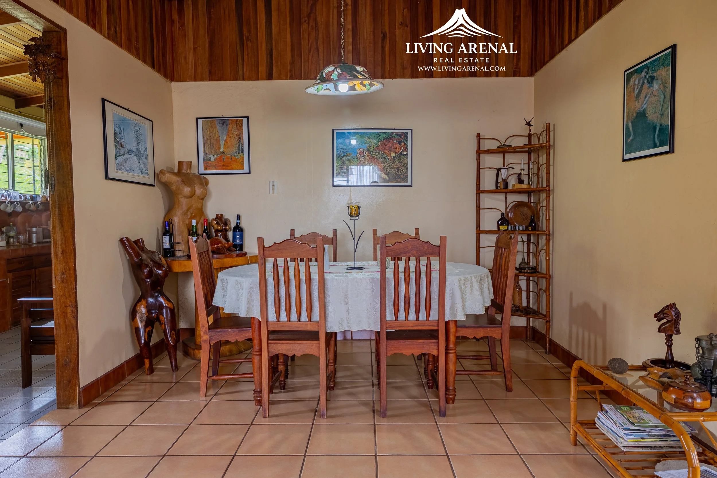 Dining room with six wooden chairs around a round table covered with a white tablecloth, decorated with a metal candle holder. Artworks hang on cream-colored walls, and there are wooden shelves with decorative items and bottles. The room has a tiled