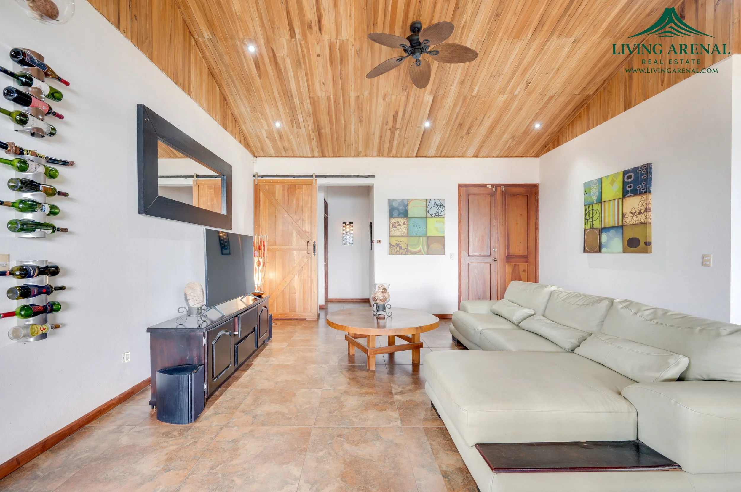 Living room with a white sectional sofa, a television on a dark wooden stand, a wooden coffee table, wine bottles on the wall, and wooden sliding barn doors. The room has a wooden ceiling with a ceiling fan and artwork on the walls.