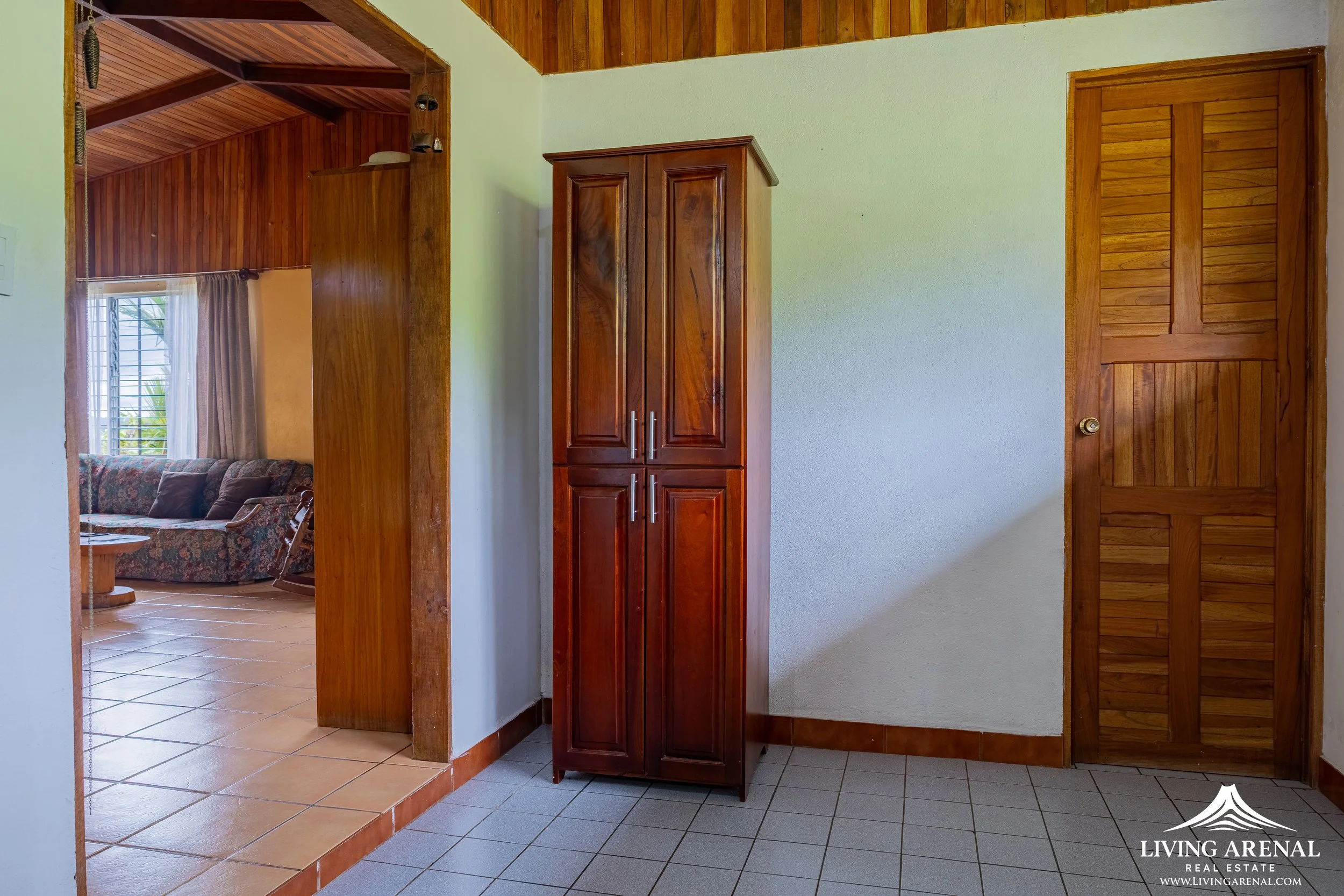 Interior of a house showing a corner with a wooden cabinet, a wooden door, and a doorway leading to a living room with a patterned sofa, a window with curtains, and tiled flooring.