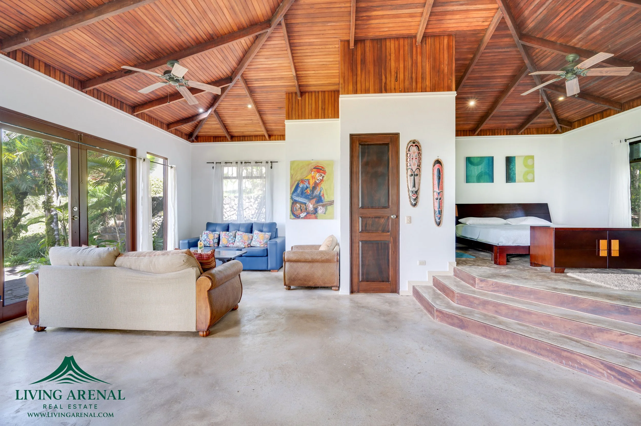 Living room with high wooden ceiling, ceiling fans, large sliding glass doors, white walls, furniture including a beige sofa, blue armchair, brown armchair, and a bed on a raised platform, with African tribal art and colorful paintings on walls, surr