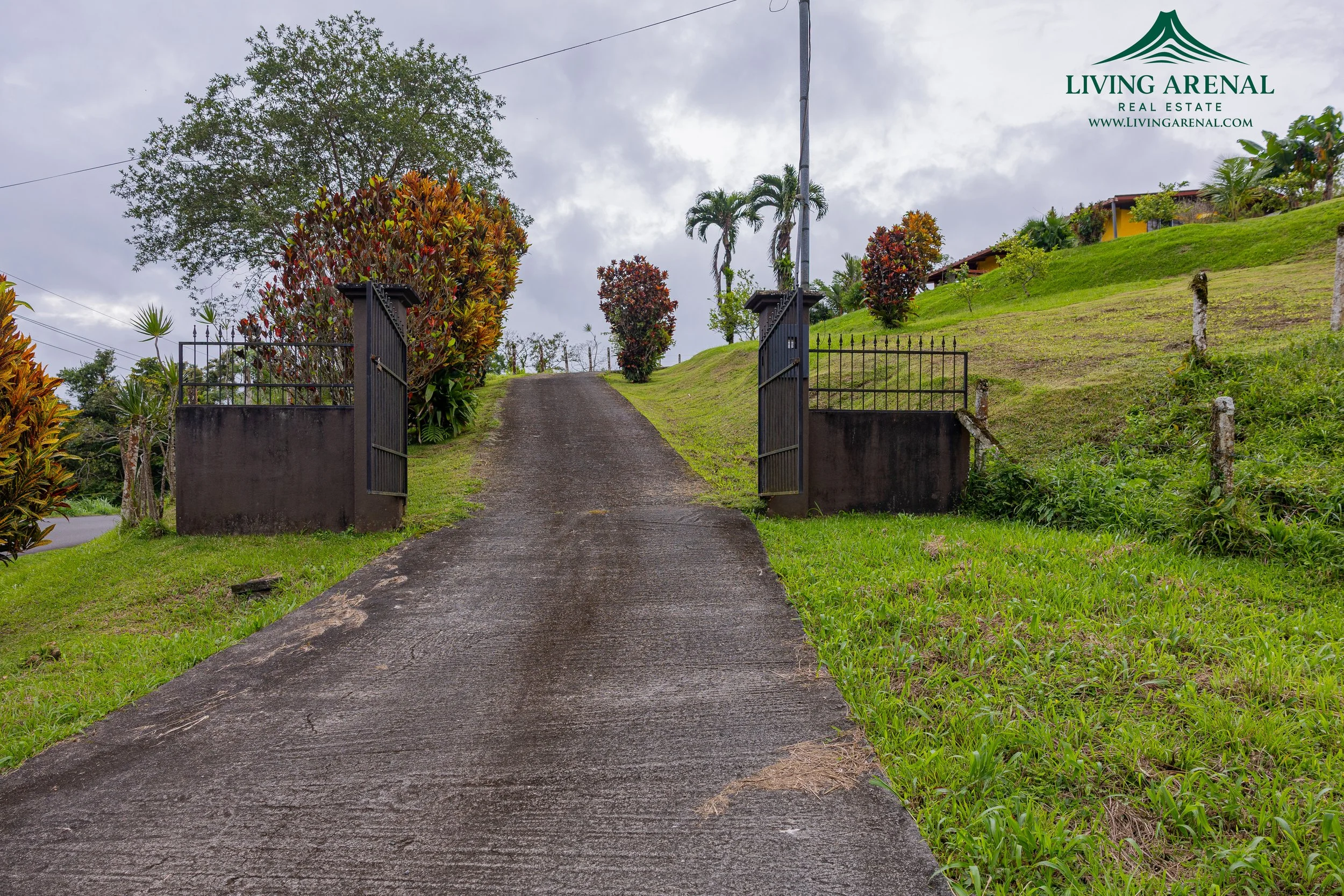 Concrete driveway leading up a grassy hill with open black iron gates on either side, surrounded by tropical trees and bushes, cloudy sky in the background, with a Living Arenal Real Estate logo in the top right corner.