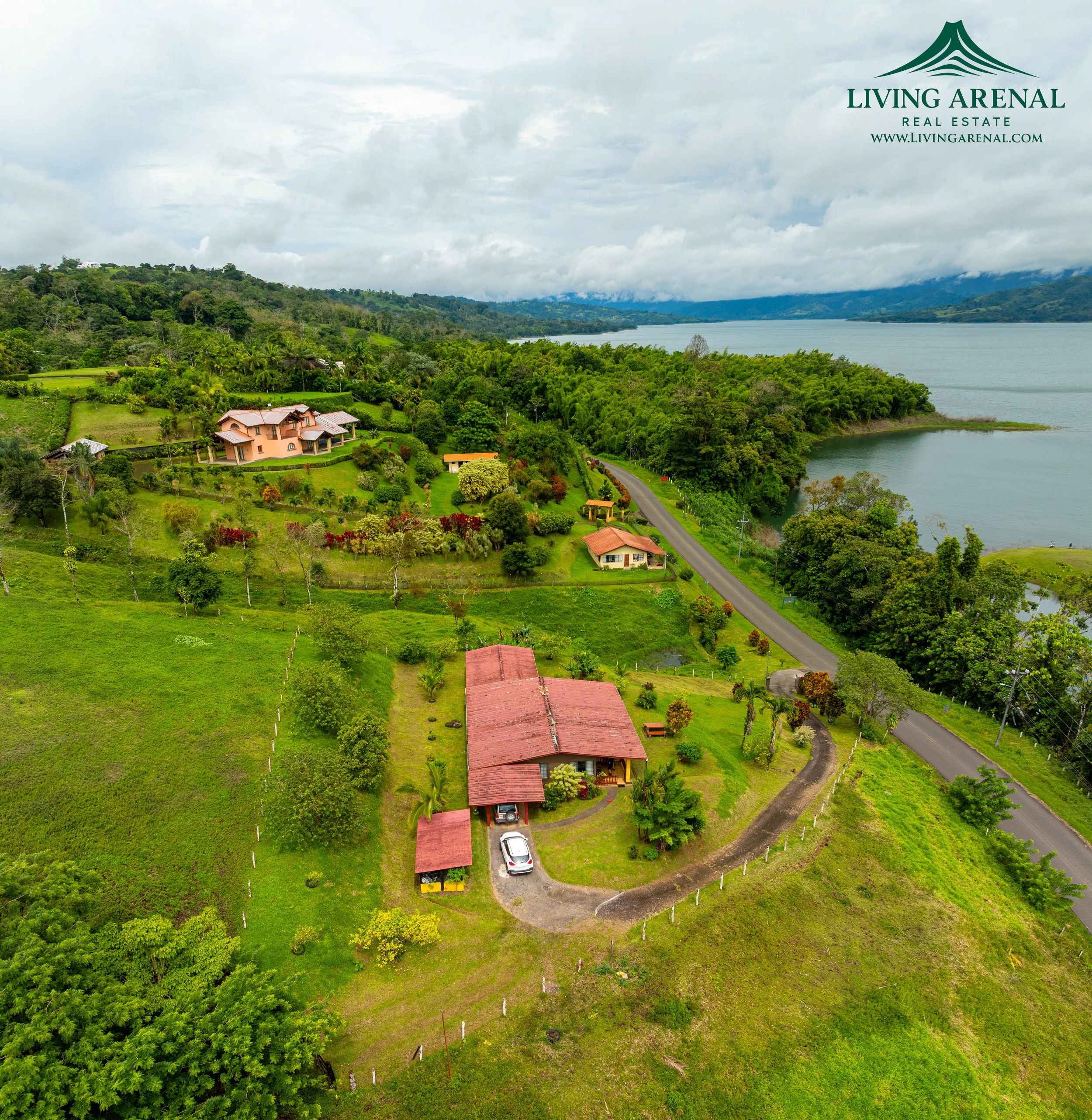 Aerial view of a lush green hillside with several houses, a winding road, and a large body of water in the background, under a cloudy sky.