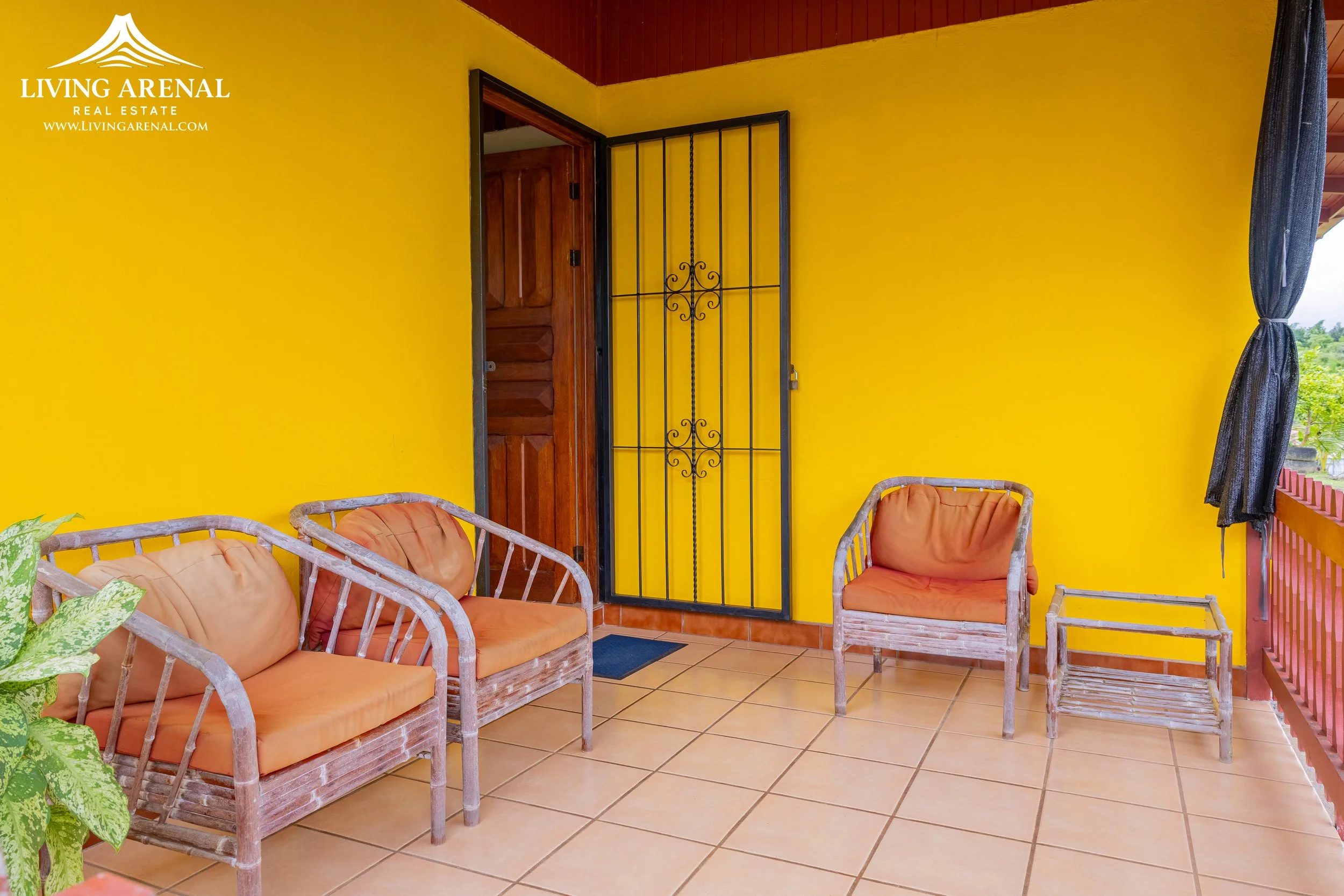 Outdoor patio with yellow walls, wooden furniture with orange cushions, a small table, and a black curtain on a door, overlooking a green landscape.