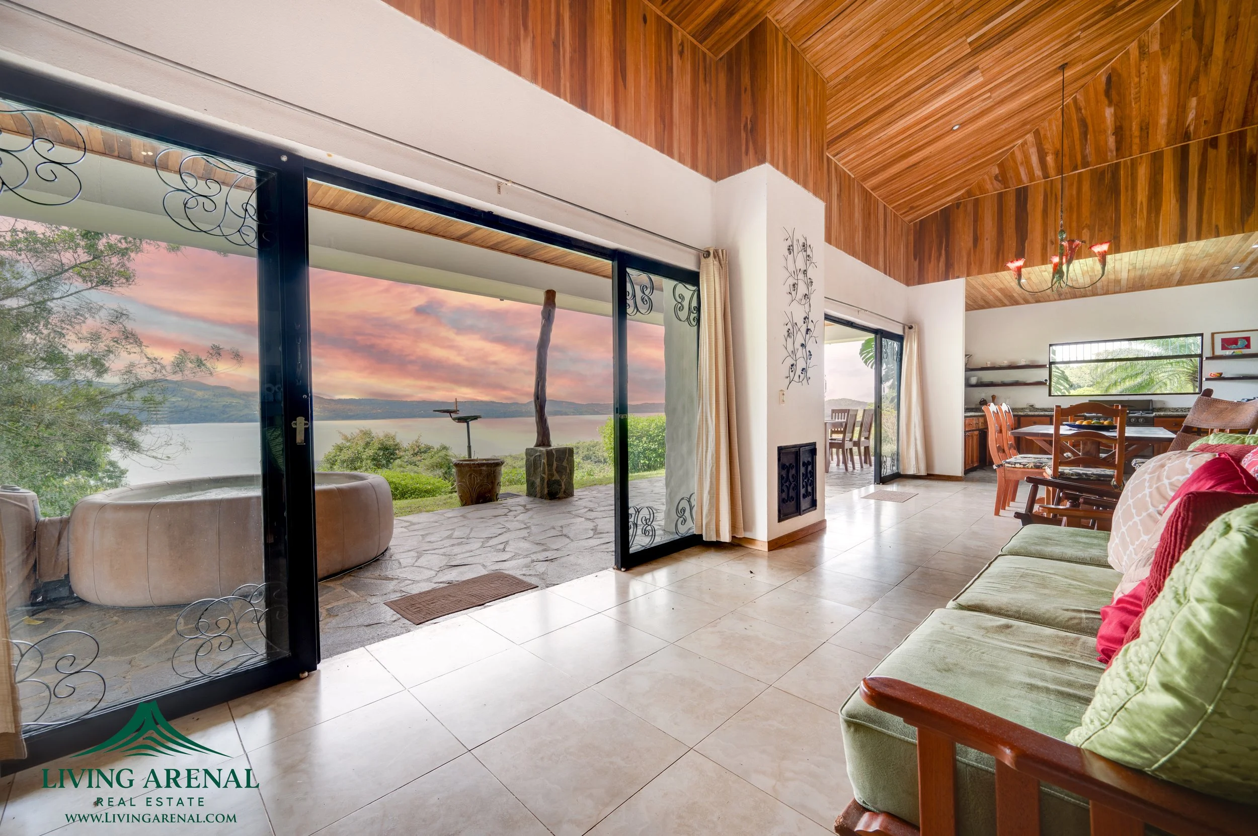 Living room with sliding glass doors open to a patio overlooking a lake and mountains at sunset. The room has high wooden ceilings, a sitting area with a green sofa, a dining table with chairs, and decorative wall art.