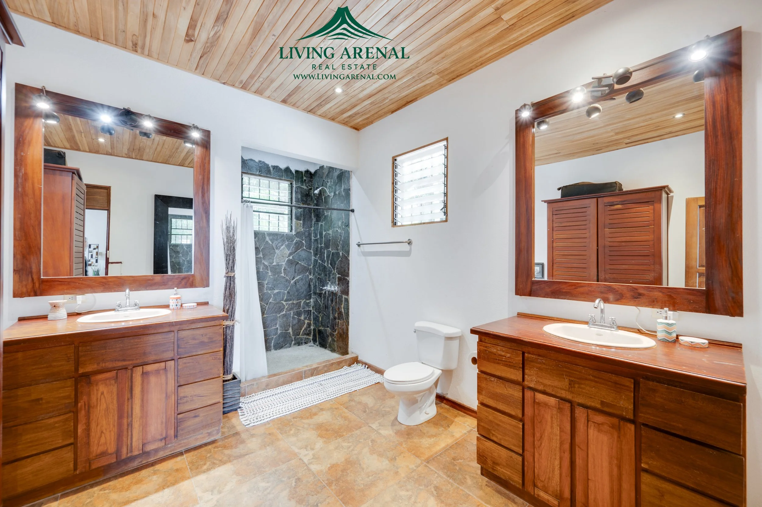 Bathroom with two wooden vanities with large mirrors, a walk-in shower with stone walls, a toilet, and wood ceiling.