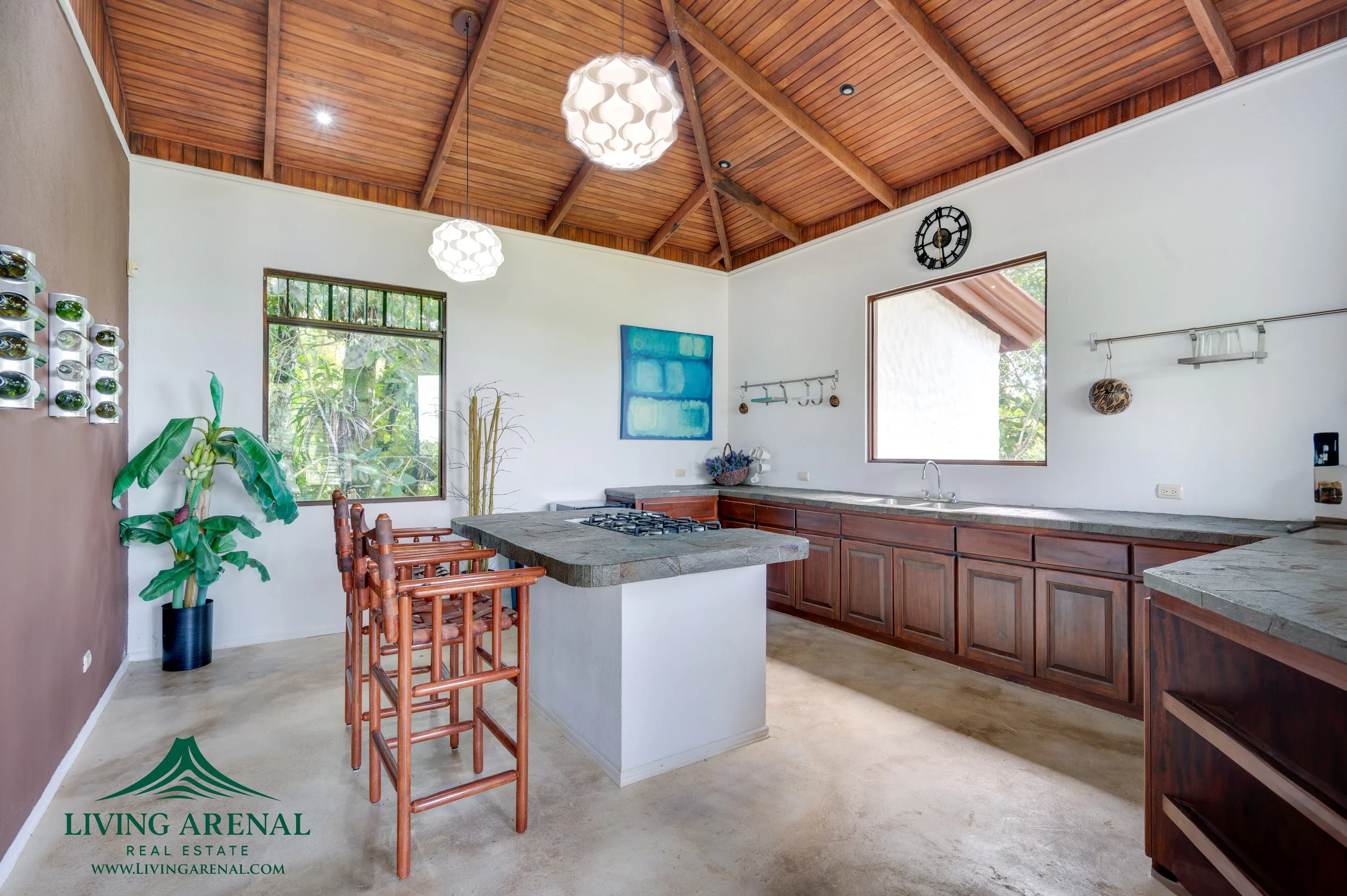Kitchen with wooden ceiling, two pendant lights, large windows, white walls, and green plant. Contains a central island with a stovetop, wooden cabinets, and a gray stone countertop. Also includes a wall clock, artwork, and kitchen tools.