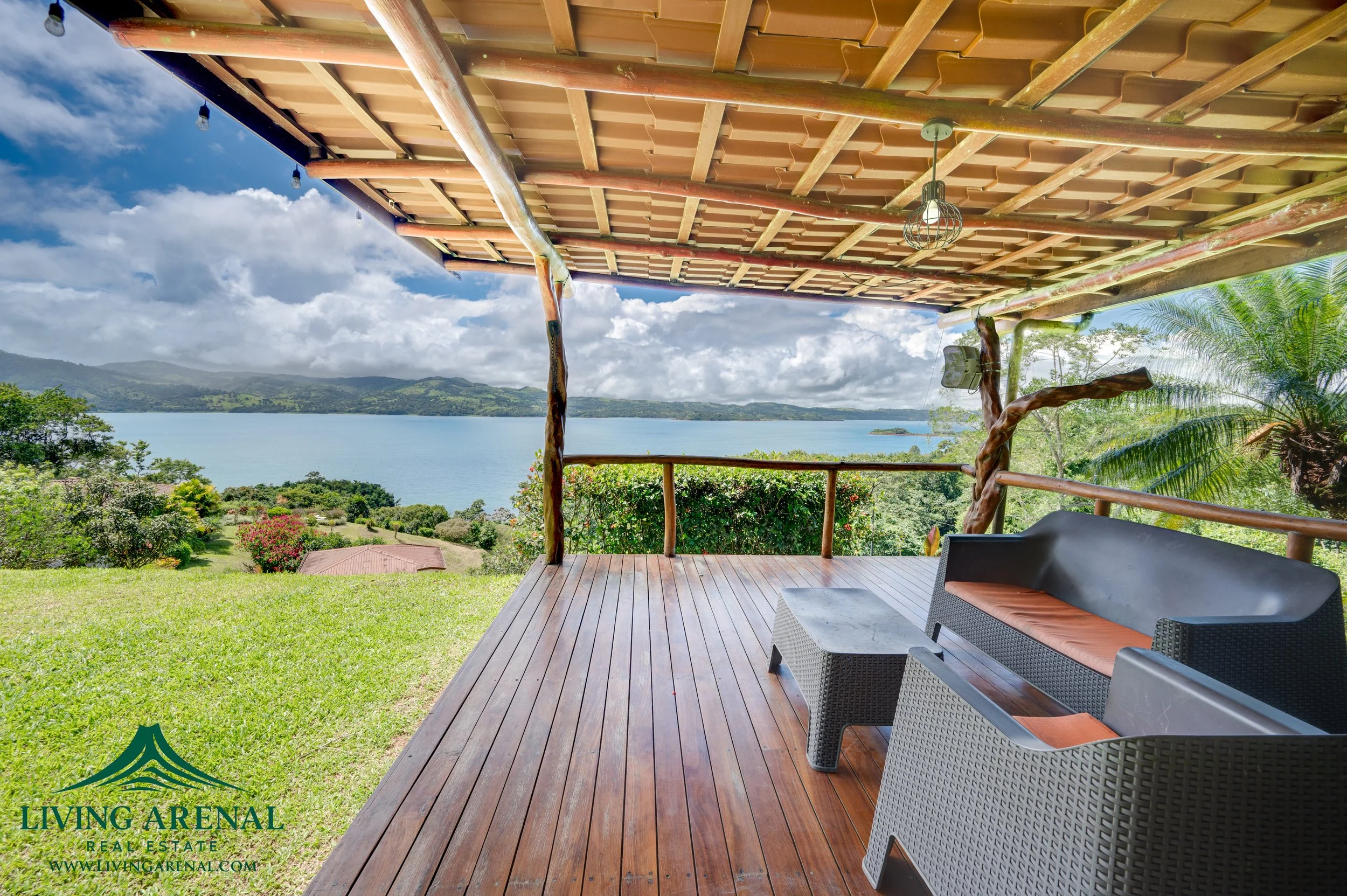 A wooden deck overlooking a lush green landscape with trees, colorful flowers, and a body of water in the distance under a partly cloudy sky.