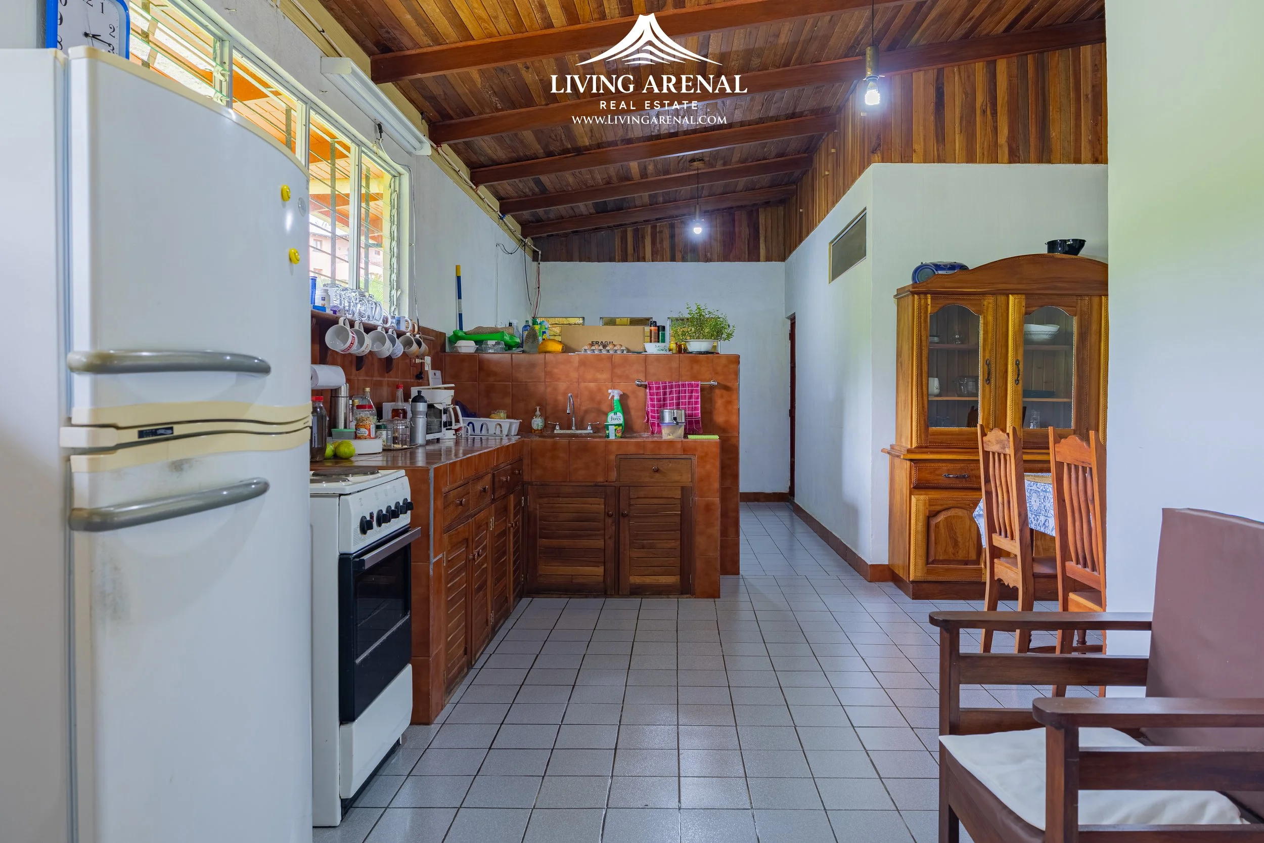 Interior of a kitchen with wooden cabinets, white refrigerator, gas stove, sink, and a seating area with chairs. There is a wooden ceiling and a tiled floor.