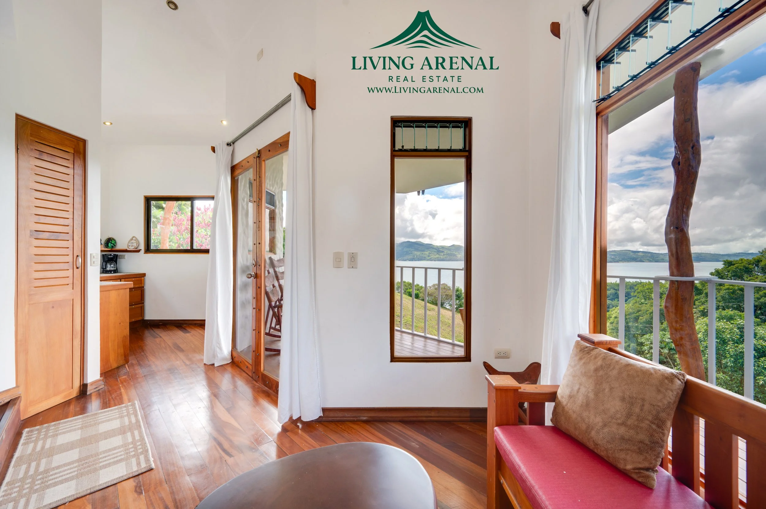 Living room with large window and door leading to balcony, showcasing scenic ocean and hillside view, wooden furniture, hardwood floors, and white curtains.