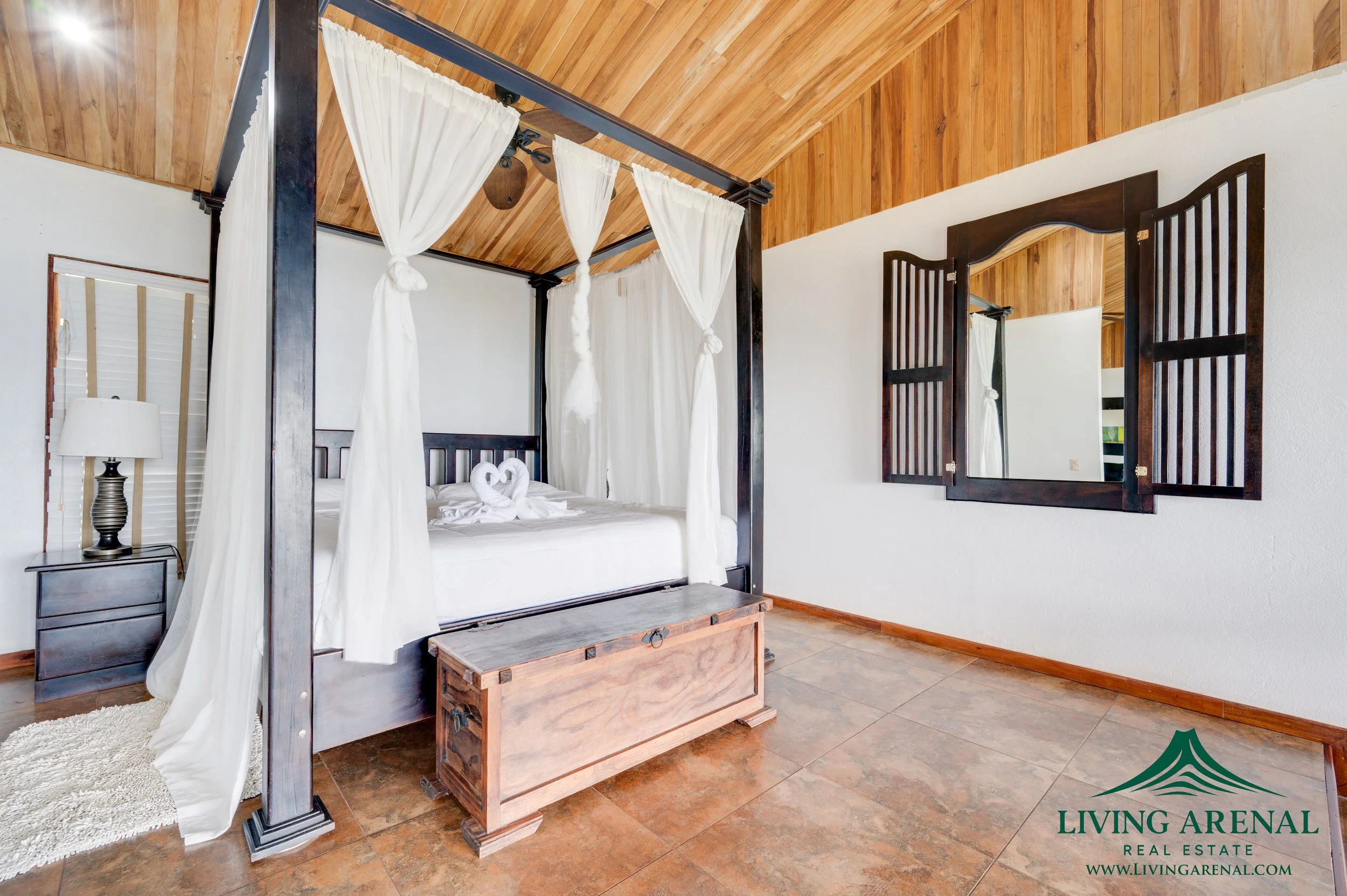 A bedroom with a four-poster bed with white curtains, a wooden chest at the foot, a nightstand with a lamp, and a wall mirror with wooden shutters, tiles on the floor, and wood-paneled ceiling.