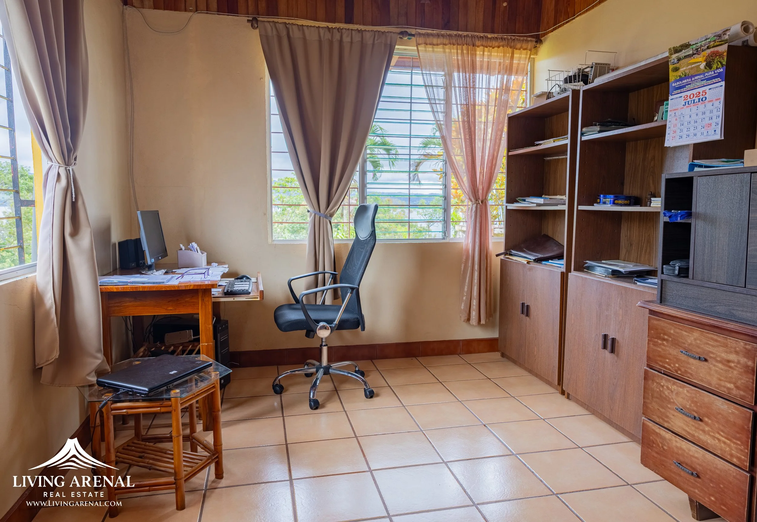 Bedroom or office with tiled floor, beige walls, and wooden shelves filled with clutter. A window with beige curtains and metal bars lets in natural light. A black office chair sits in front of a desk with a computer, phone, and paperwork. There's a