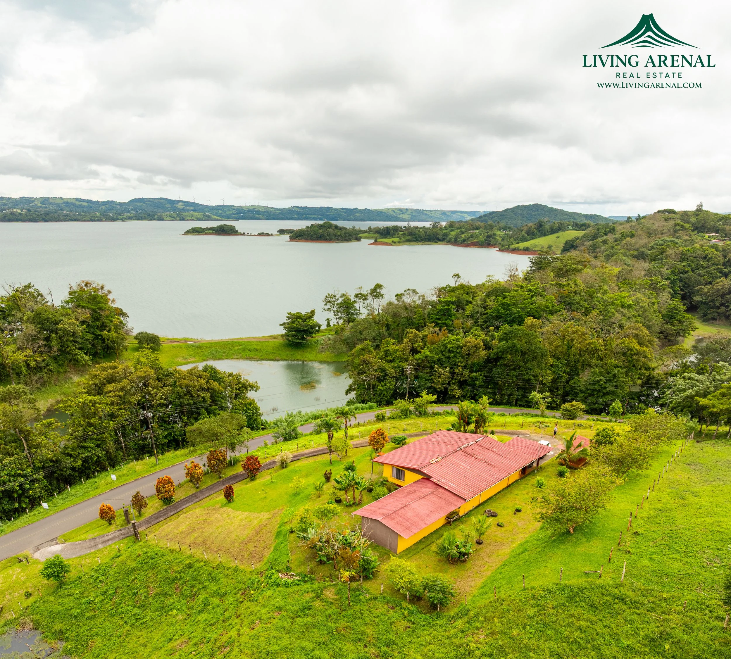 Aerial view of a yellow house with red roof surrounded by greenery and trees, overlooking a lake with hills and islands in the distance, partly cloudy sky.