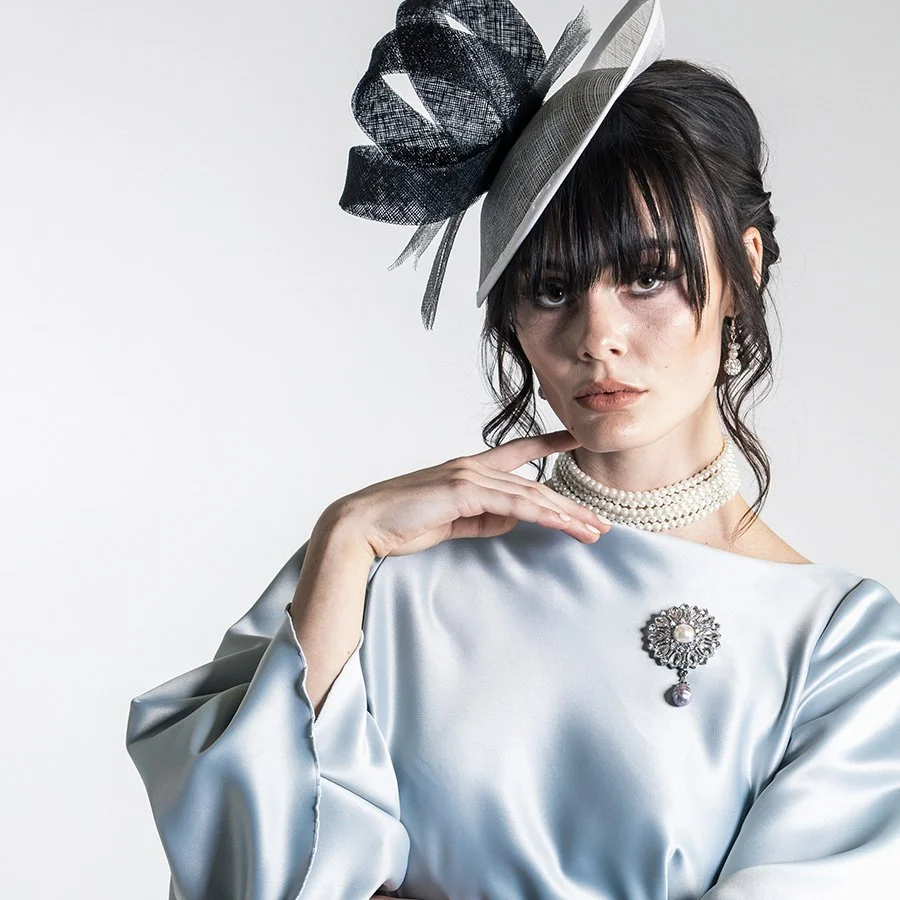 A woman wearing a shiny silver satin with a pearl choker and earrings, and a decorative brooch on her dress. She has dark hair with bangs and wears a stylish hat with black and white netting. She has a serious expression and is touching her neck with her right hand.