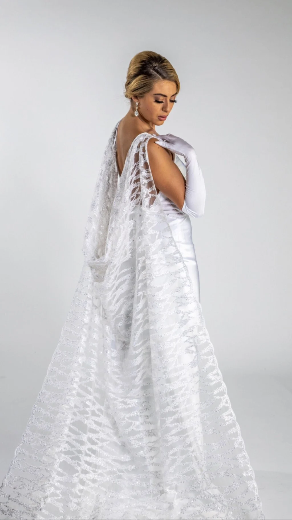 A woman wearing a white wedding dress and gloves, with a lace veil, posing against a plain background.