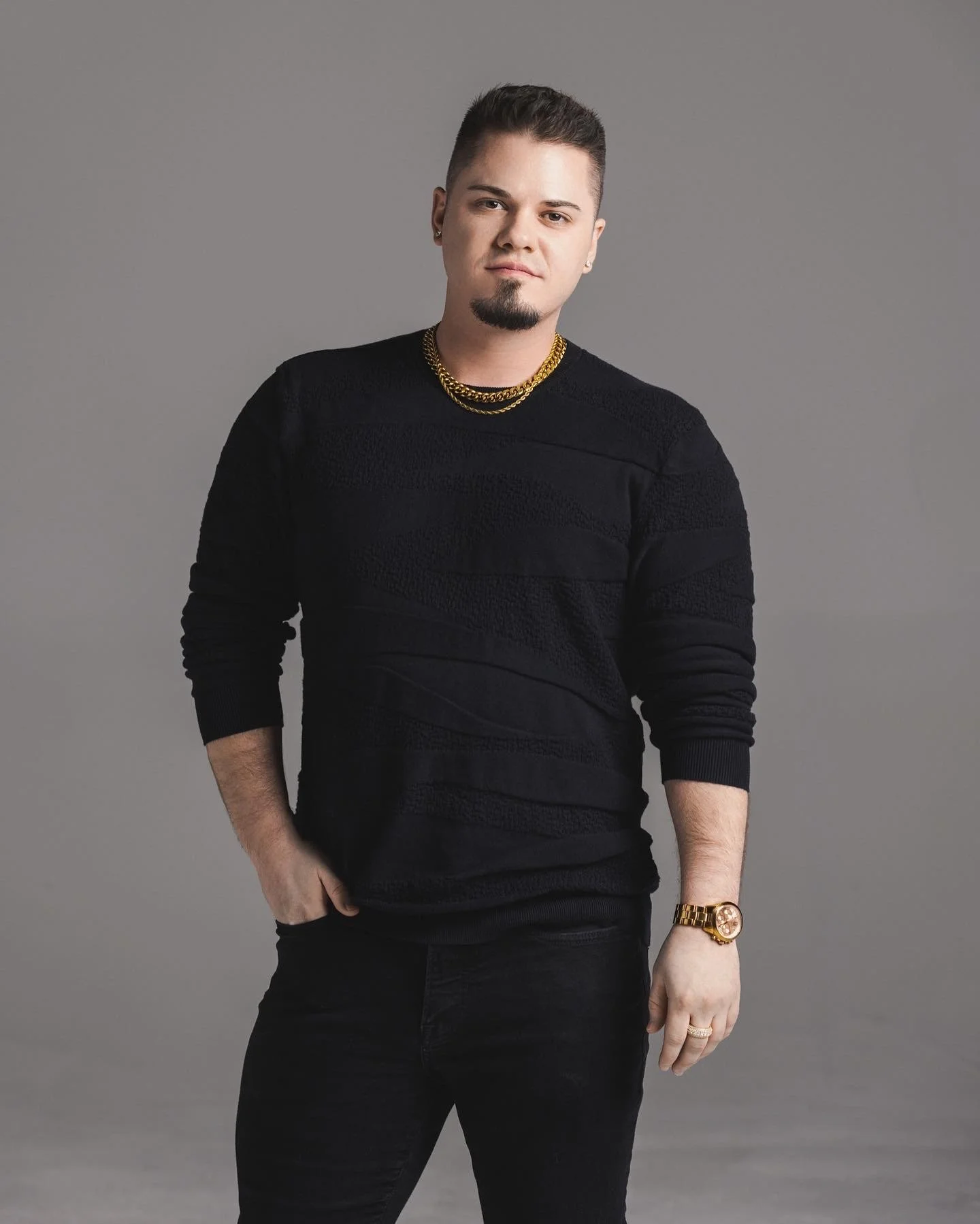 A young man with short dark hair, a goatee, and earrings wearing a black sweater with textured stripes, black pants, gold jewelry including a chain necklace, bracelet, and watch, standing against a gray background.