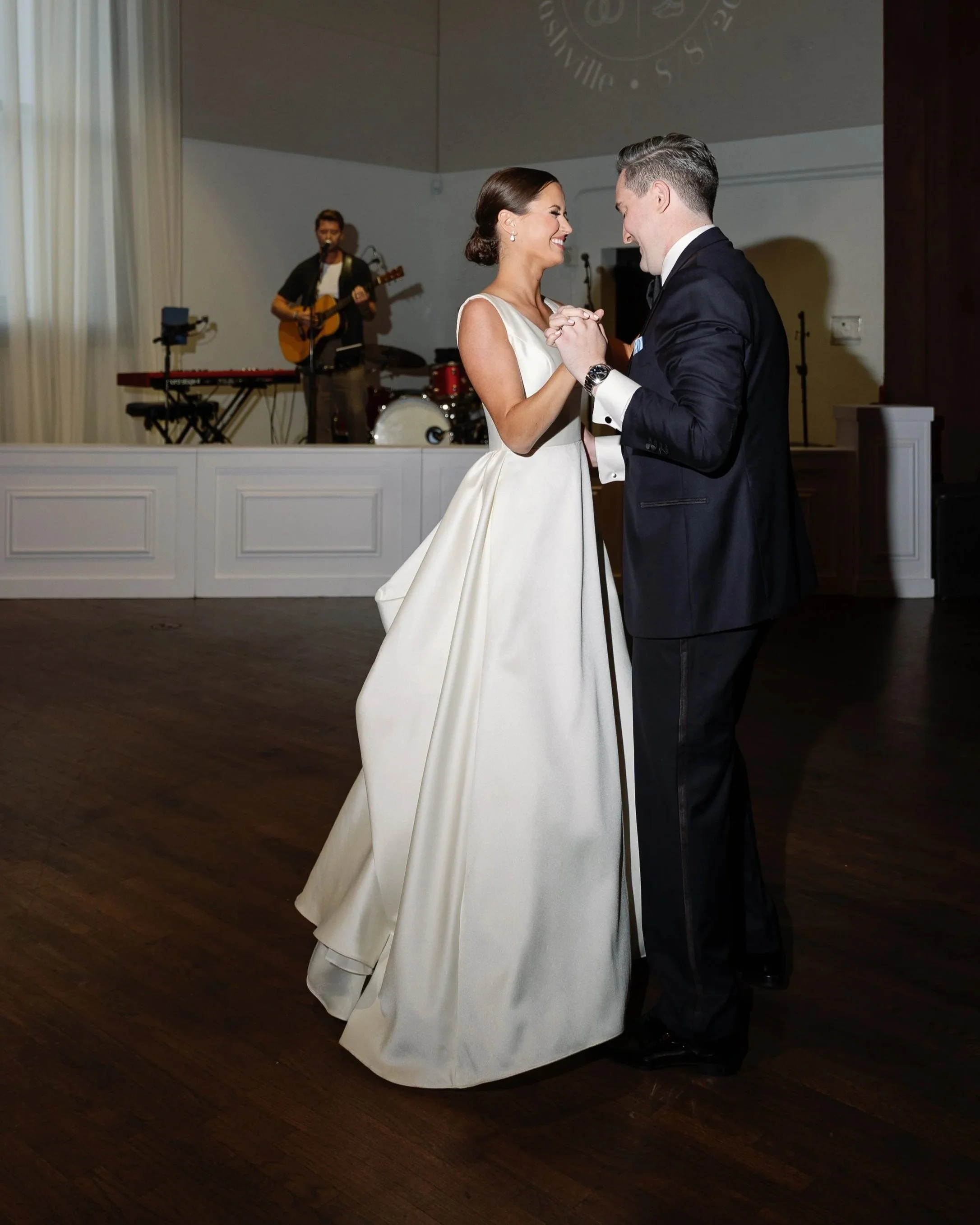 A bride and groom dancing together at their wedding reception, with a musician playing guitar and singing in the background.