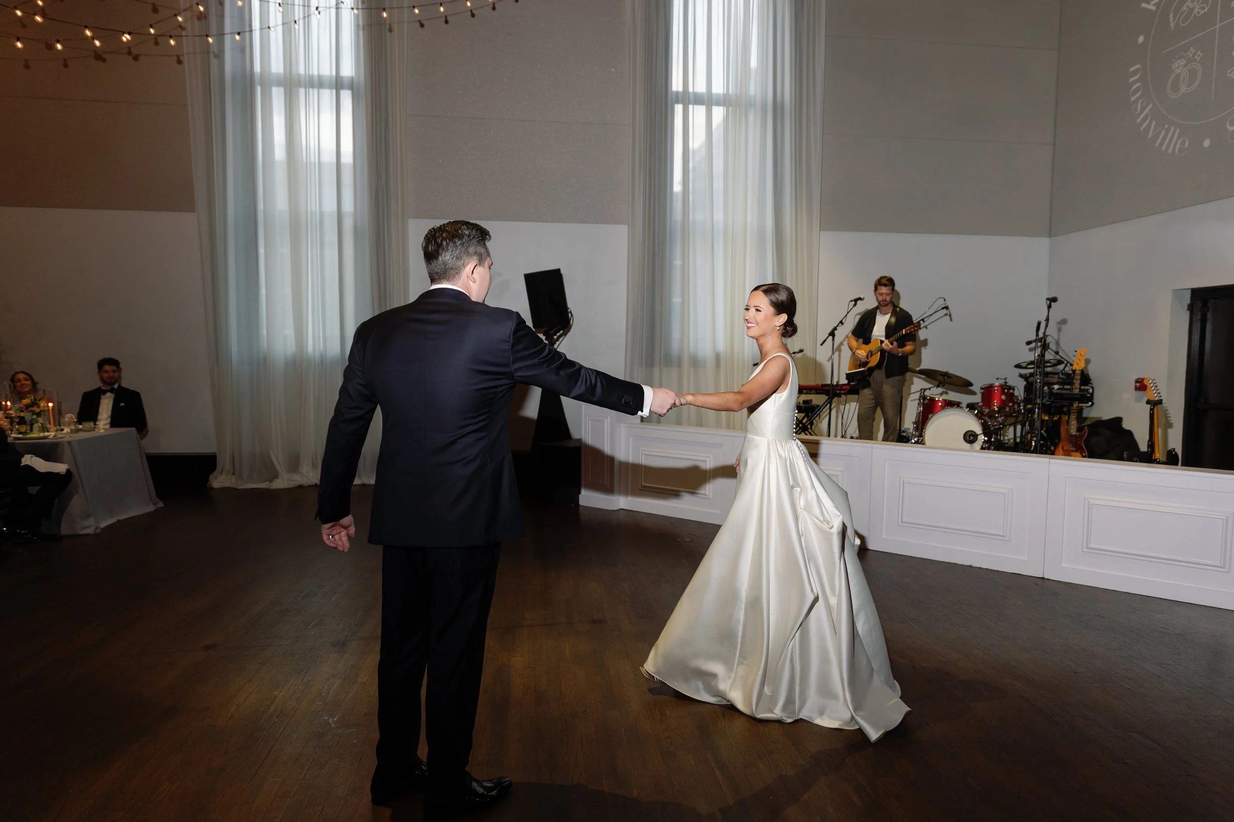 A bride and groom dancing at their wedding reception, with a live band playing in the background and guests seated at decorated tables.