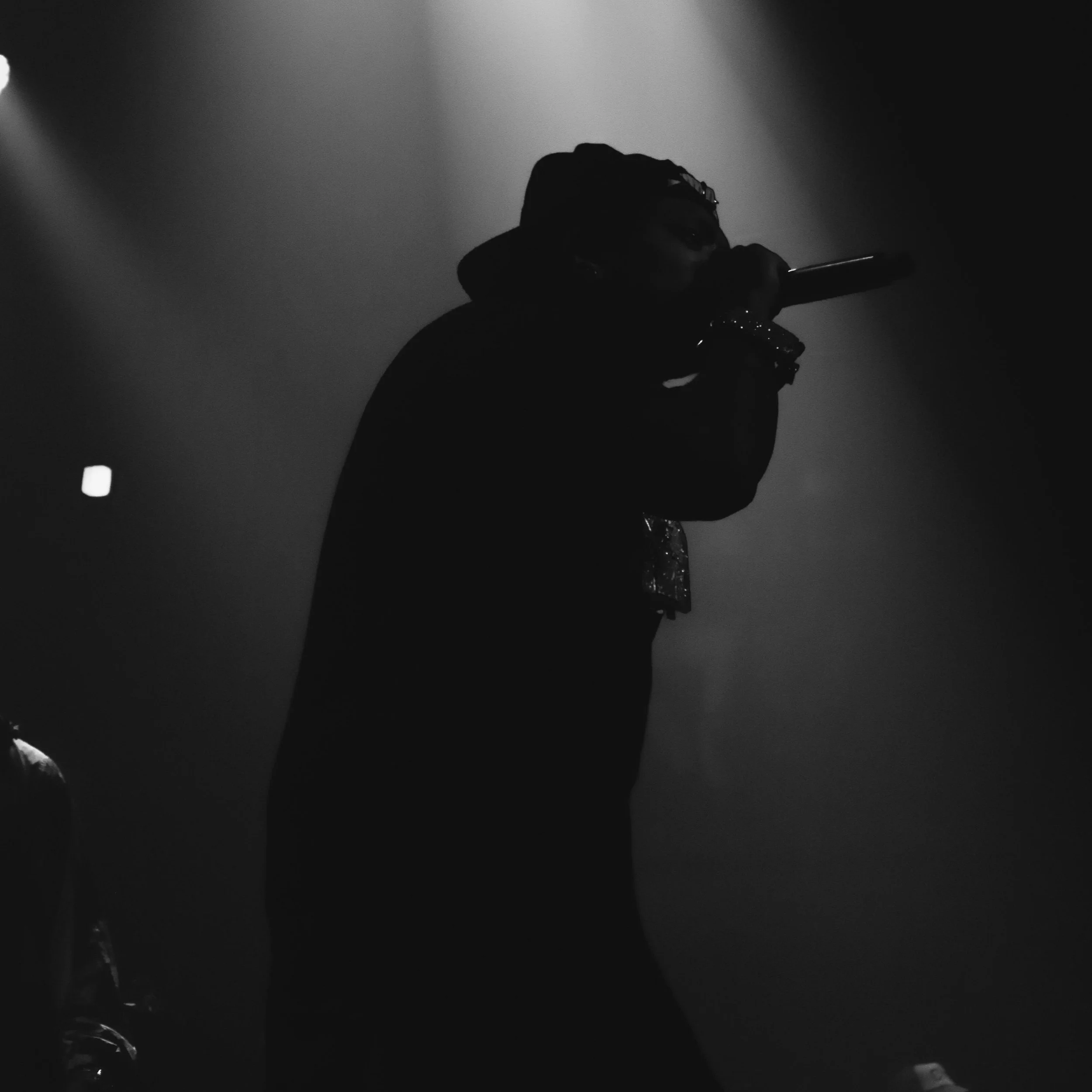 A silhouette of a person wearing a cap and jewelry, holding a microphone, performing on stage in low light.