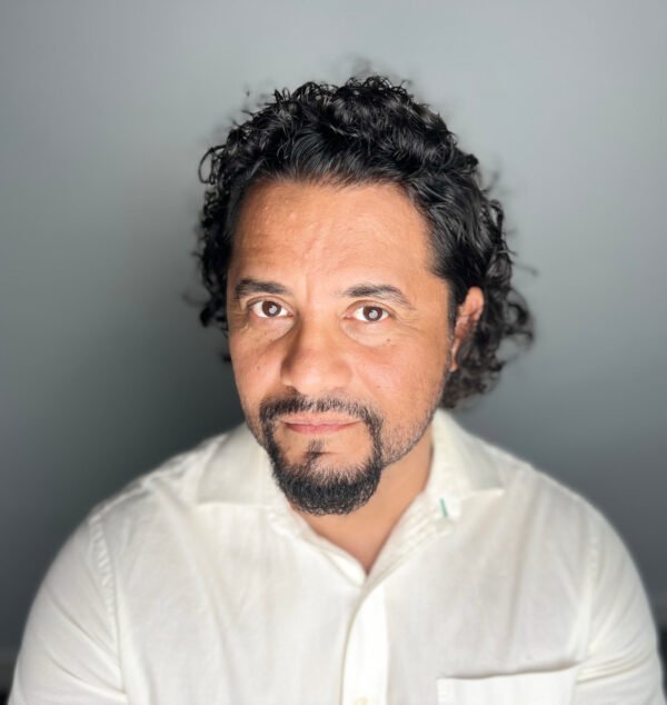 Portrait of a man with dark curly hair and a goatee, wearing a white button-up shirt, against a plain gray background.