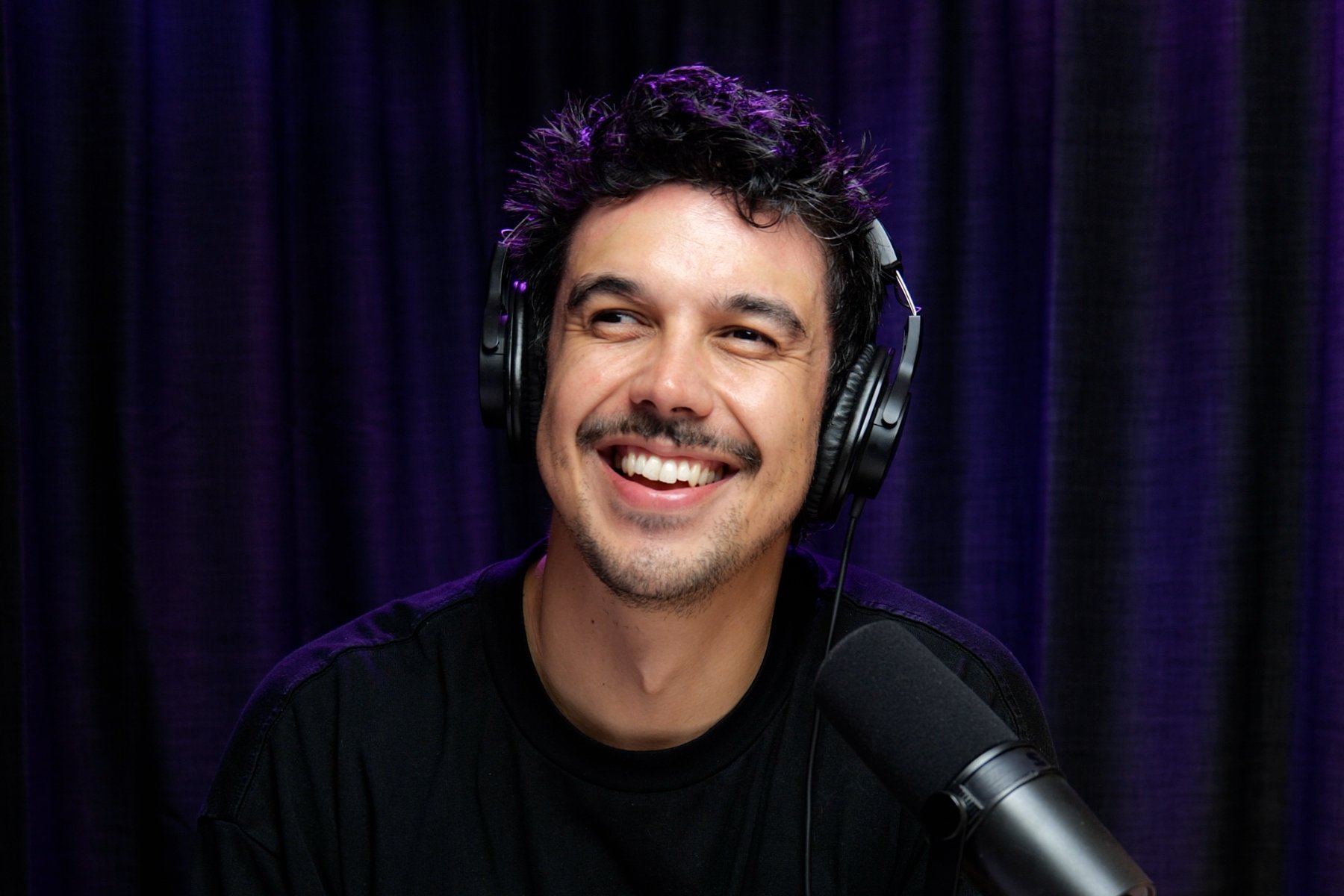 A man with curly dark hair and a beard, smiling while wearing headphones and speaking into a microphone in a studio with dark purple curtains.
