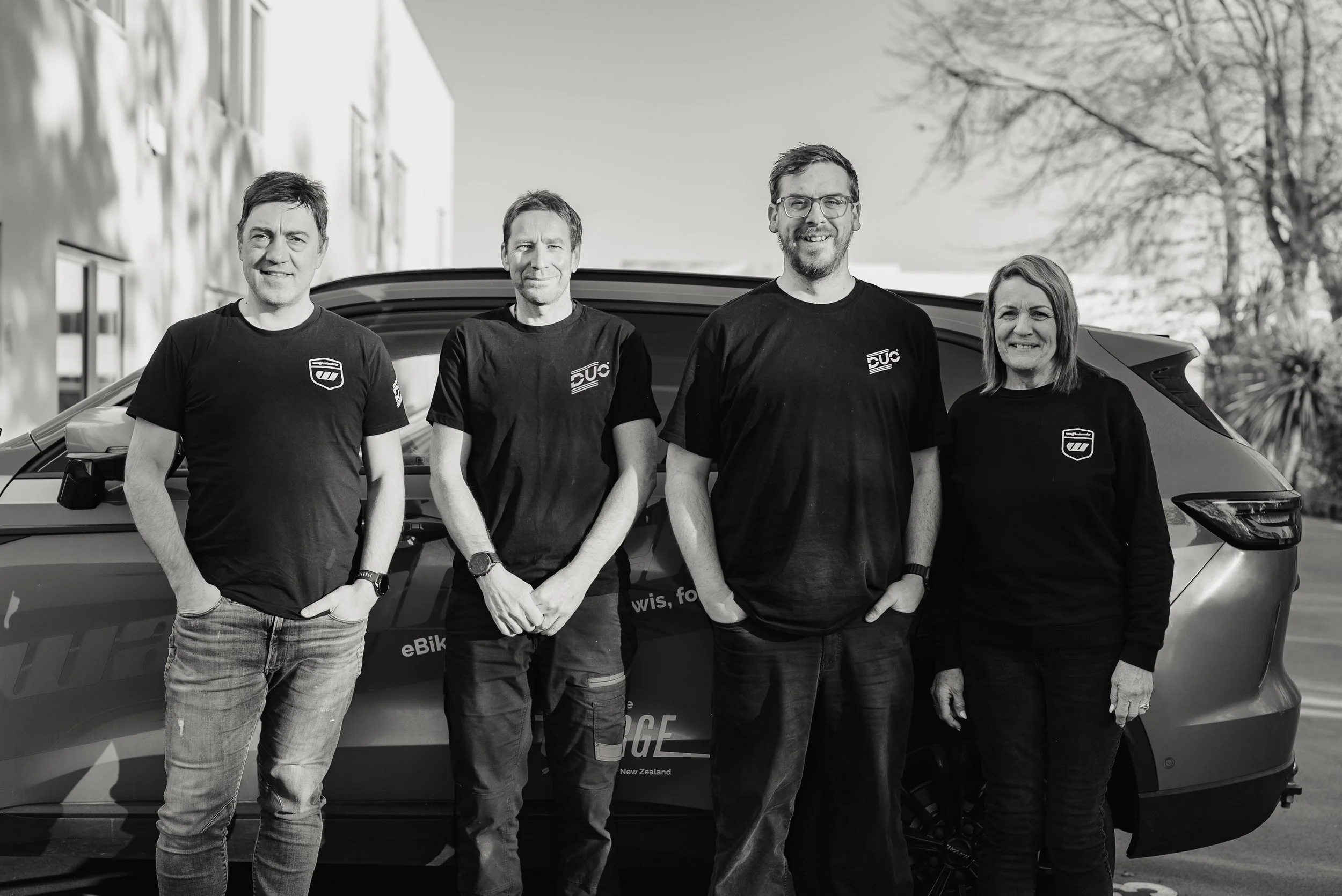Four people standing in front of a car outdoors, smiling at the camera. The background includes trees and a building.