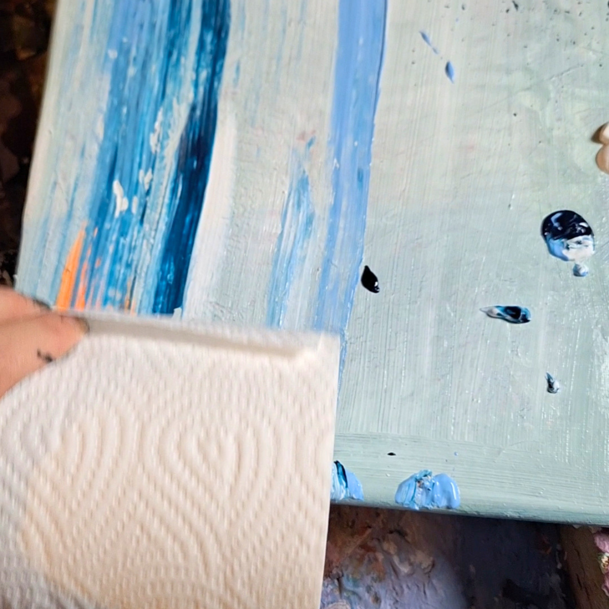 Close-up of a person wiping a painted canvas with a paper towel. The canvas has textured strokes of blue, white, and hints of orange paint.
