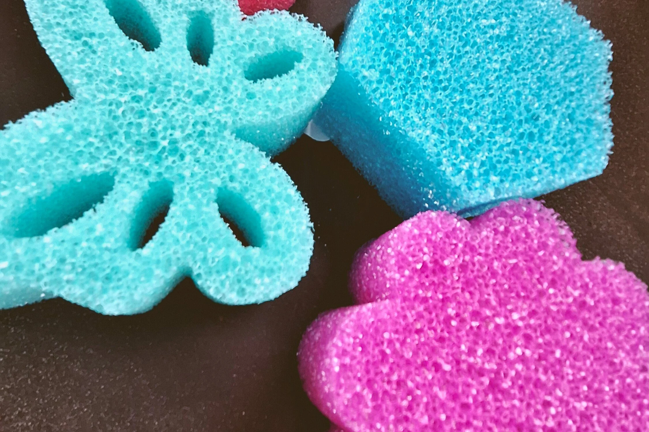 Colorful cleaning sponges, including a pink heart-shaped sponge, a bluish-green flower-shaped sponge, and a blue rectangular sponge, on a dark surface.