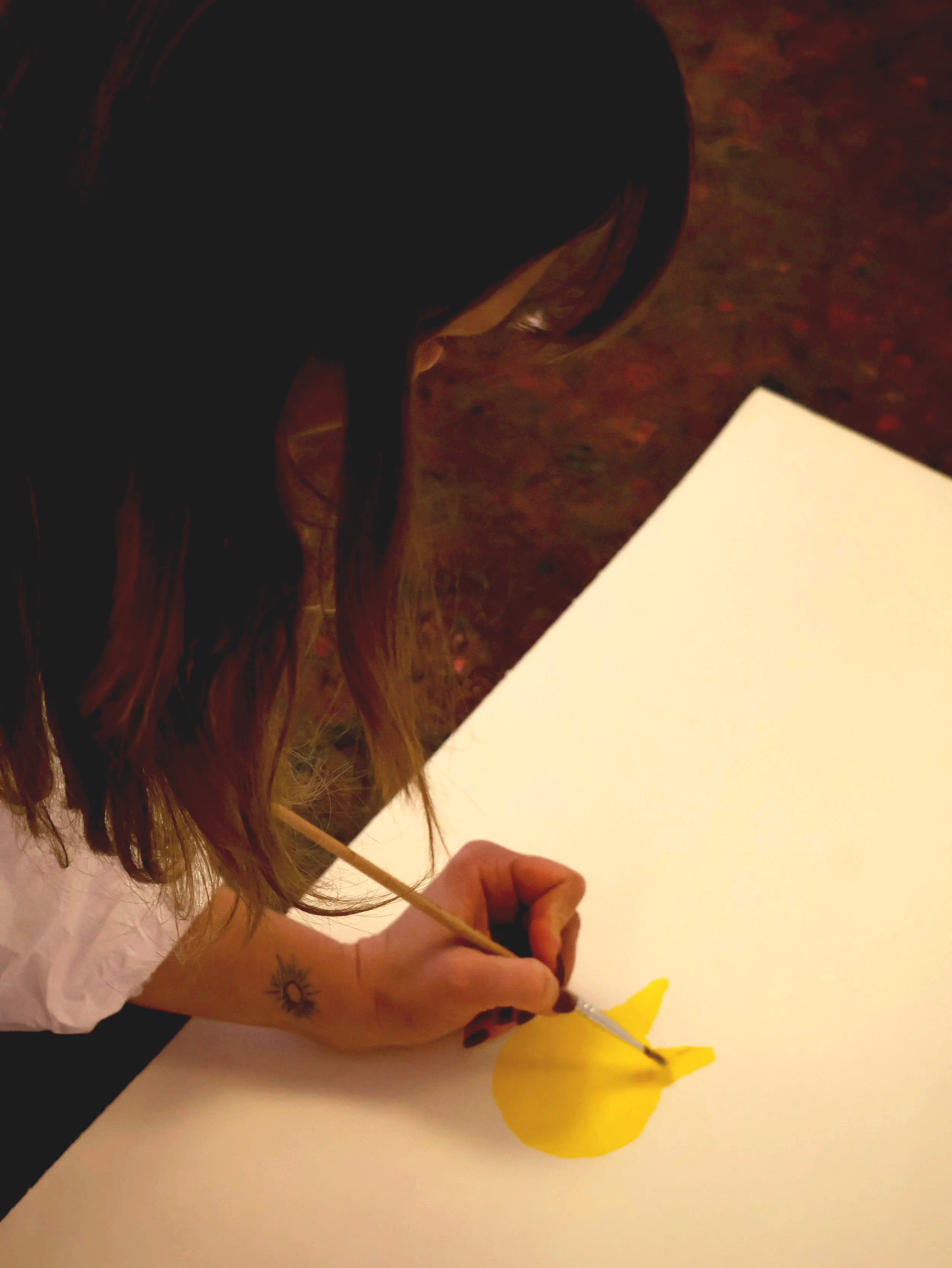 A person with long hair is coloring paper with yellow paint, creating a yellow shape on a white sheet of paper. The person has a small tattoo of a sun on their left forearm.