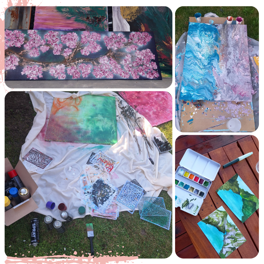 Multiple paintings and art supplies displayed outdoors. One painting features pink cherry blossoms on a dark background, another with swirling blue and purple patterns, and a third with a landscape of green trees and blue sky. Various brushes, paints, and canvases are arranged around the artworks.