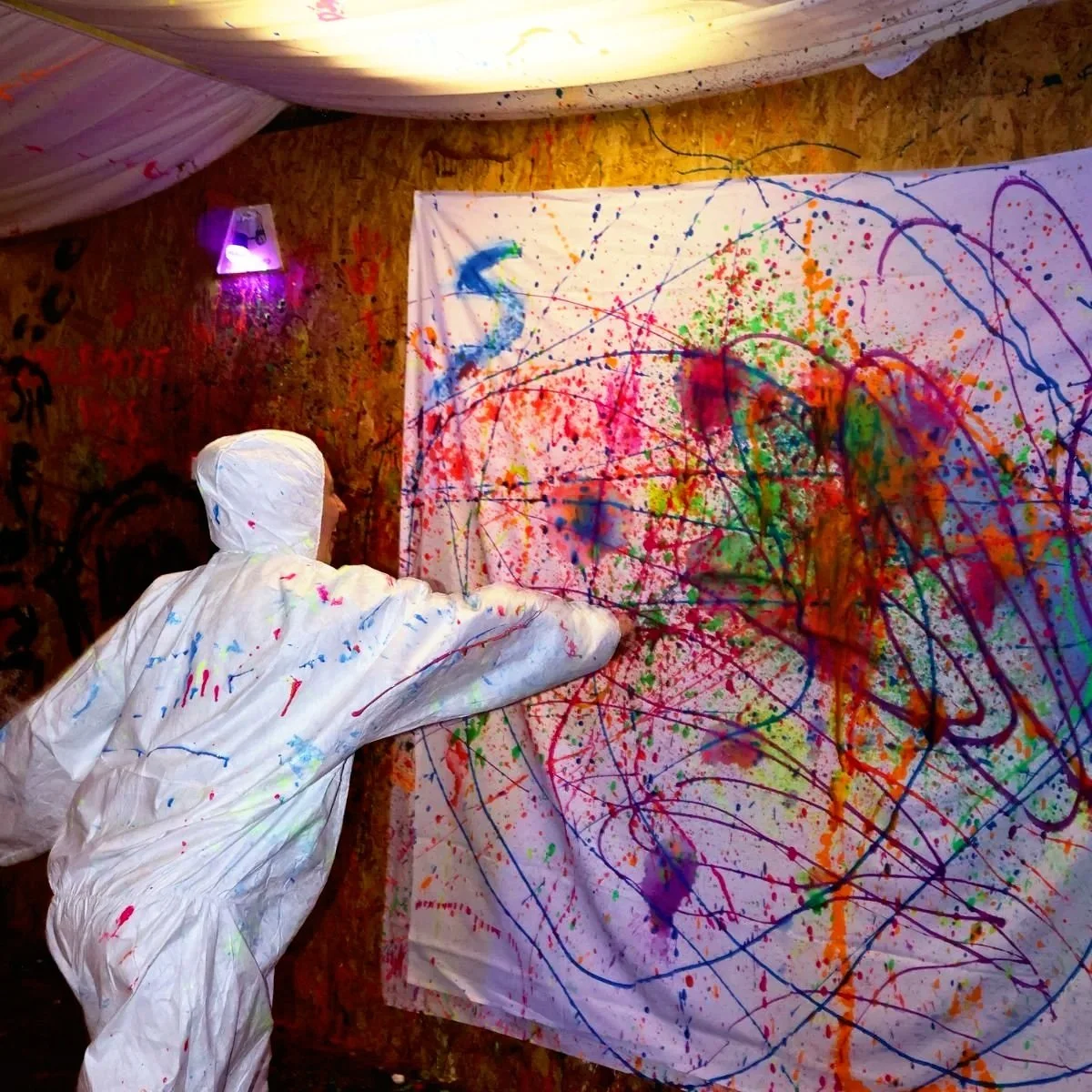 A person in a white protective suit is painting abstract splatters and strokes of various bright colors on a large canvas with spray and brush techniques, set in a room with dim lighting and textured wooden walls.