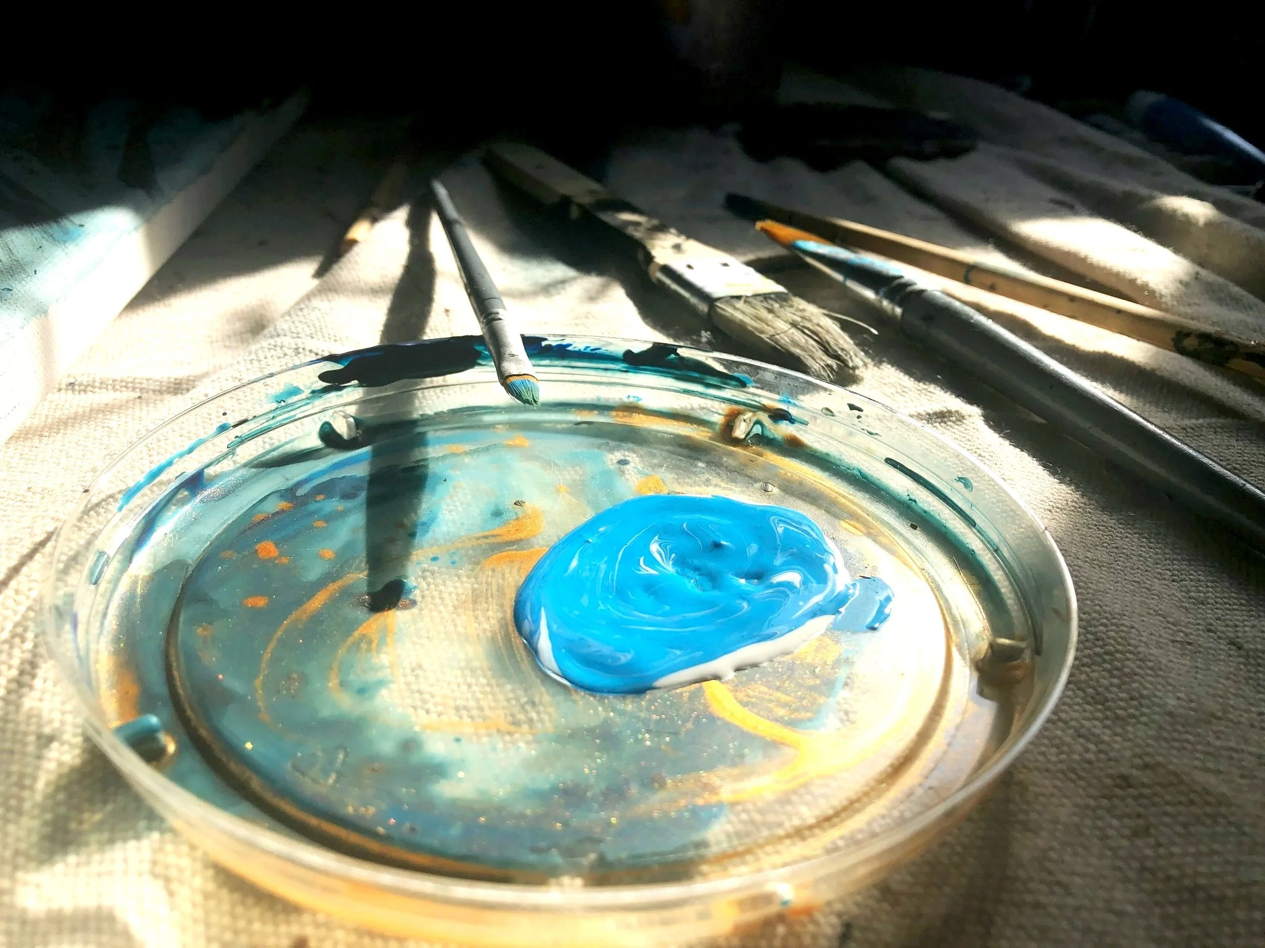 Close-up of a small container filled with blue paint, with several paintbrushes and a paint scraper resting on a textured fabric surface.