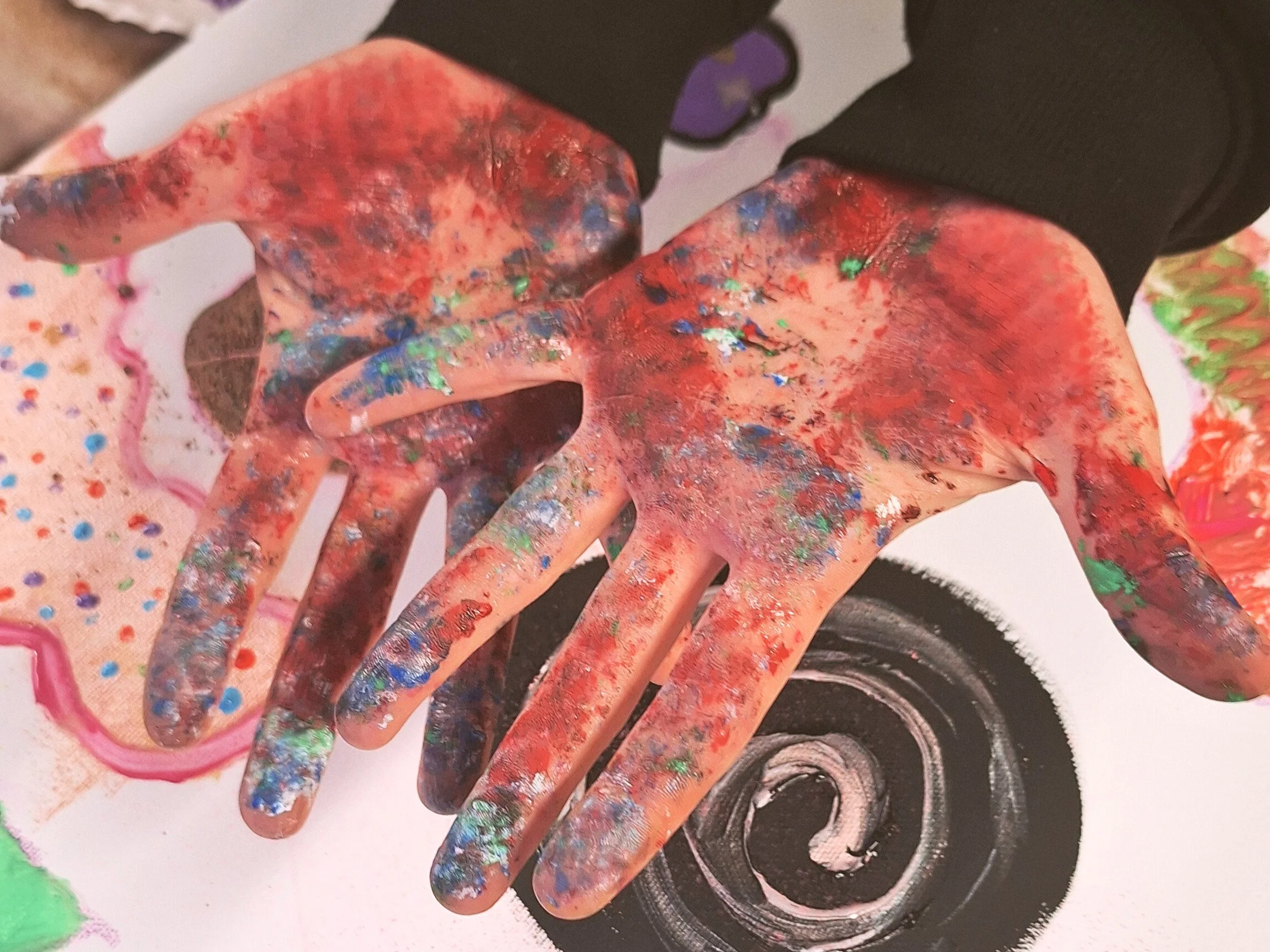 A person's hands covered in multicolored paint, with a black sleeve on one arm, creating abstract artwork with a mix of colors, including red, blue, green, silver, and pink.