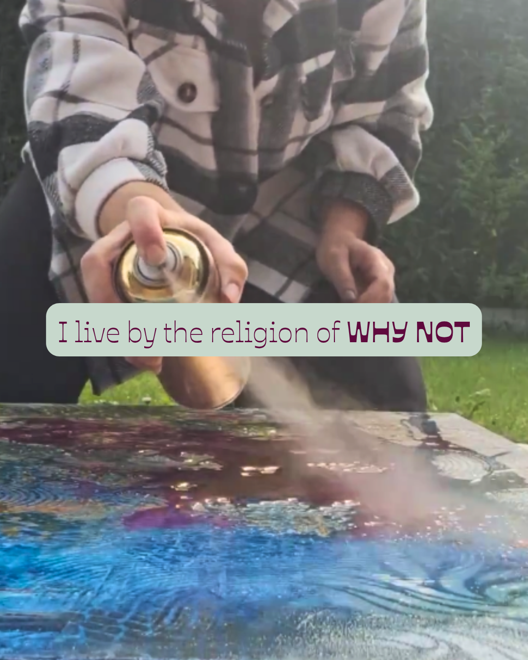 Close-up of a person in a plaid shirt using a spray can on a large, colorful, painted surface outdoors, with text overlay stating "I live by the religion of WHY NOT."
