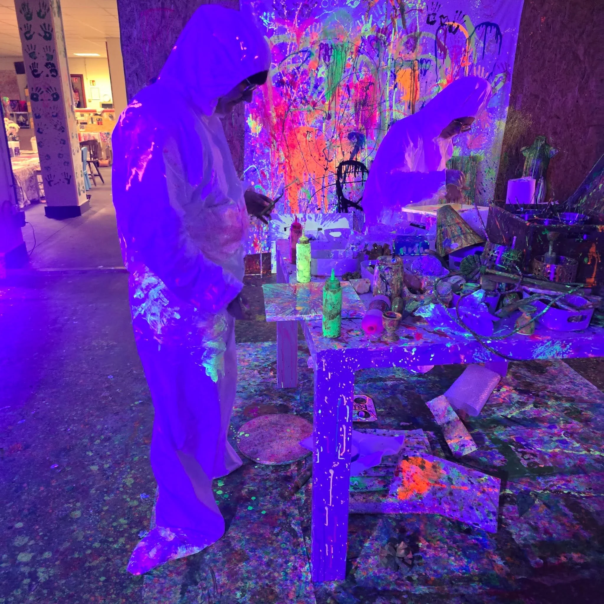 Individuals in protective suits creating glow-in-the-dark or fluorescent art with paint on a wall and objects in a dark room with blacklight, surrounded by colorful splatters and handprints.