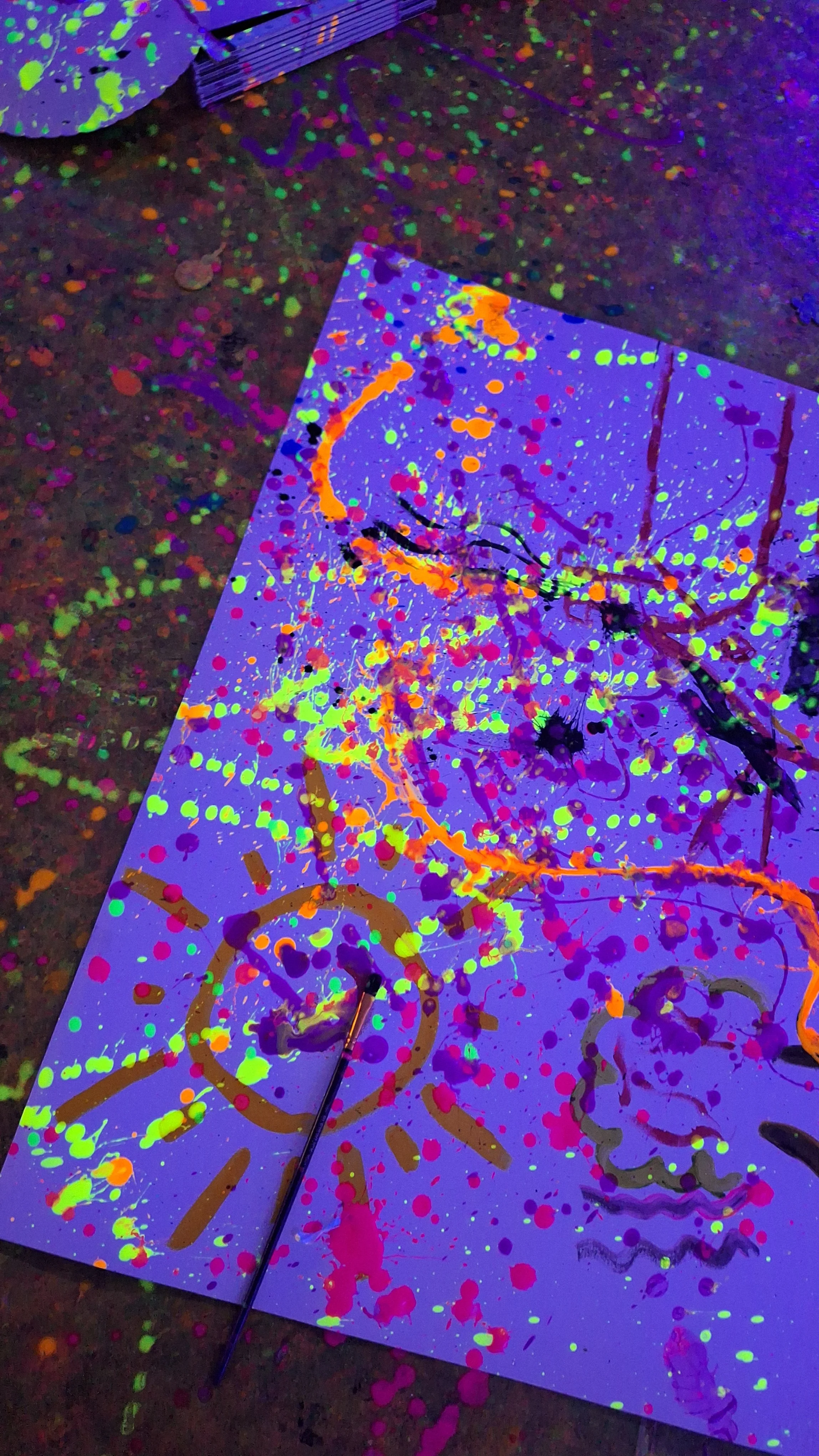 Colorful splatters and abstract patterns under black light on a surface, with some paintbrushes visible.