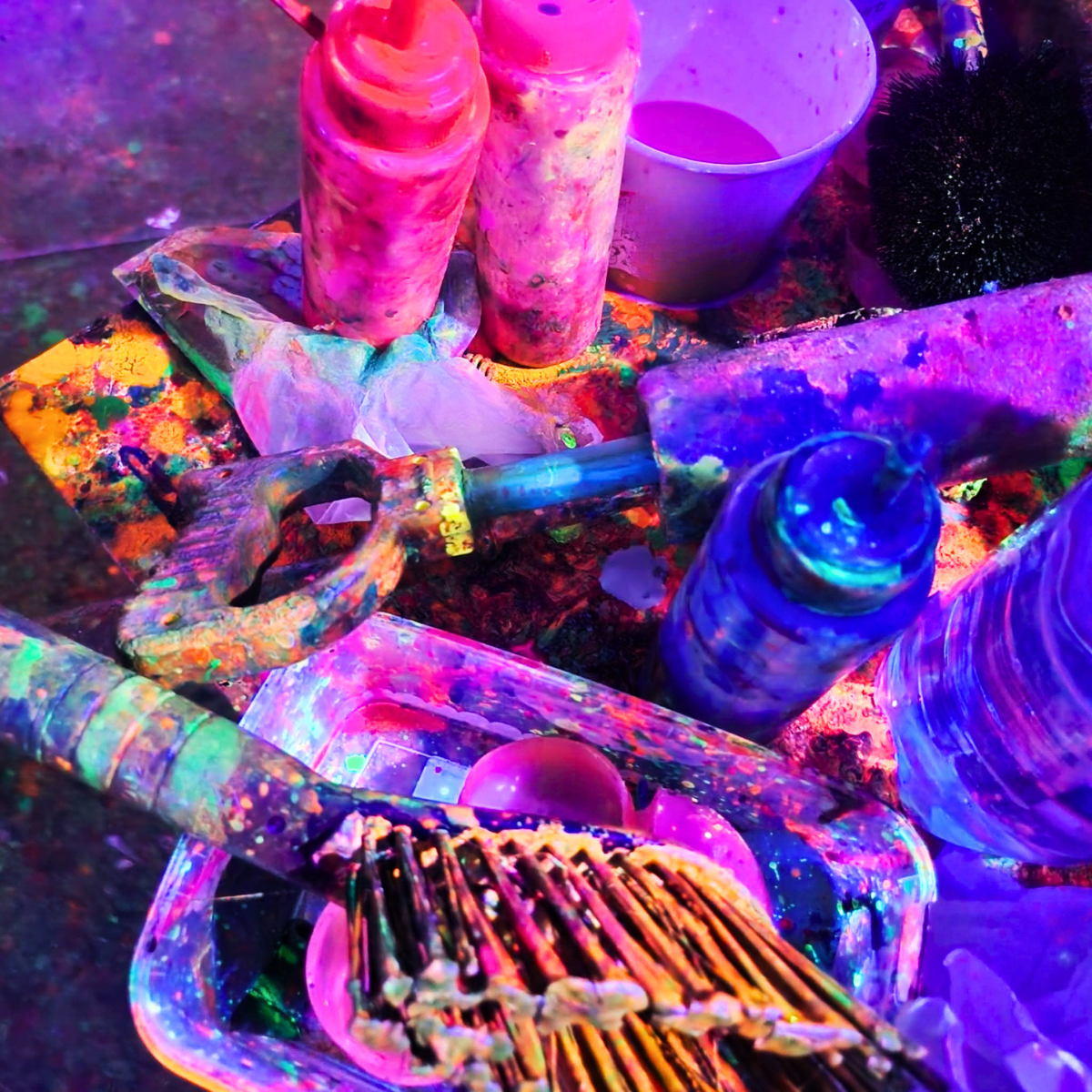 Close-up view of a colorful, abstract art setup with various objects, including bottles, a hammer, a cup, a wicker basket, and other items, all covered in vibrant paint under UV light.