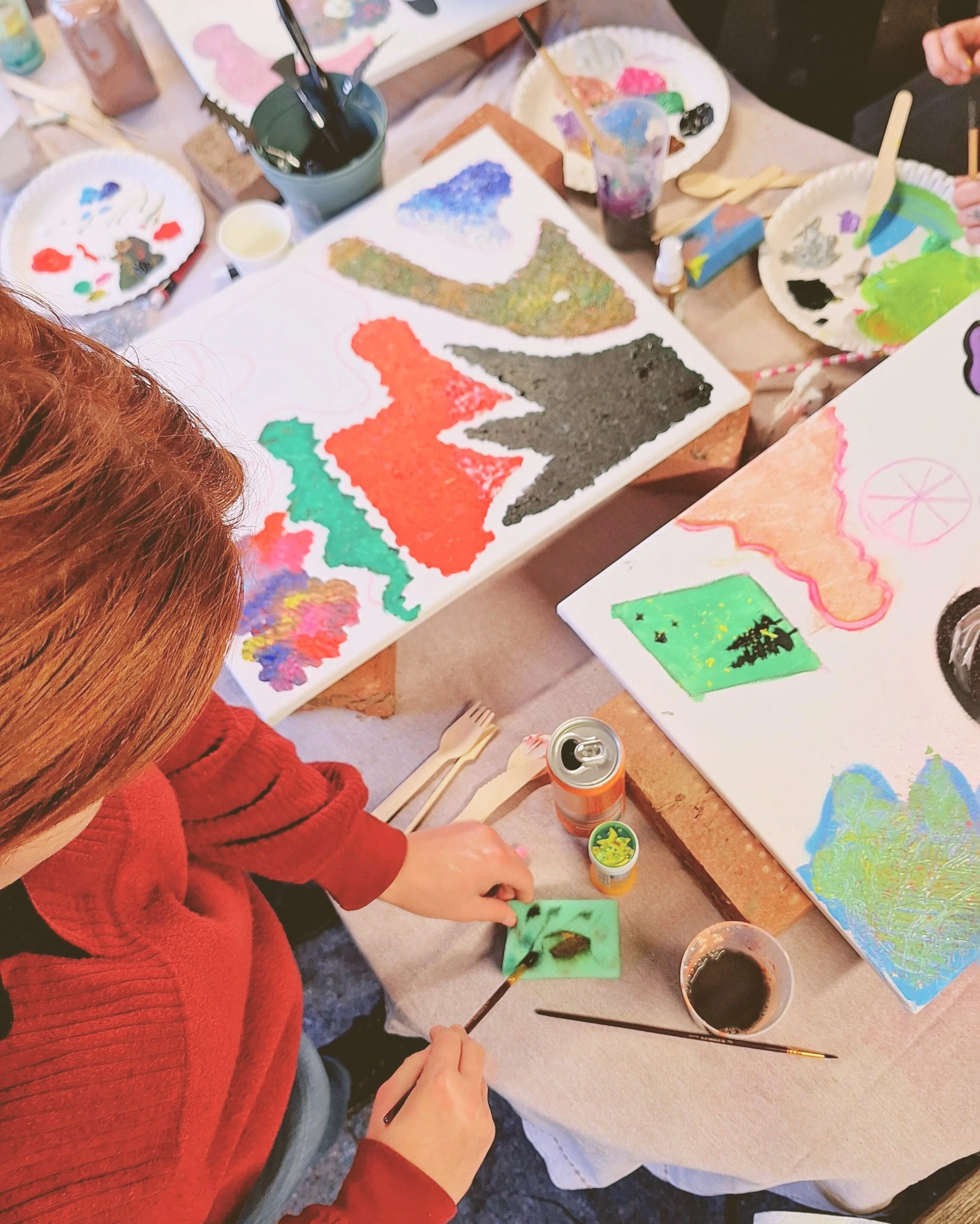 Person with red hair and red sweater painting abstract artwork with various colors and textures on canvases, surrounded by painting supplies and brushes.