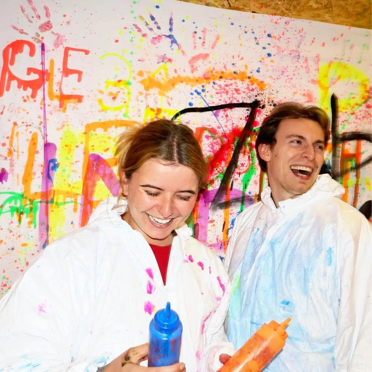 Two smiling people in white paint-splattered coveralls in front of a colorful, splattered wall holding paint bottles, celebrating a colorful event.