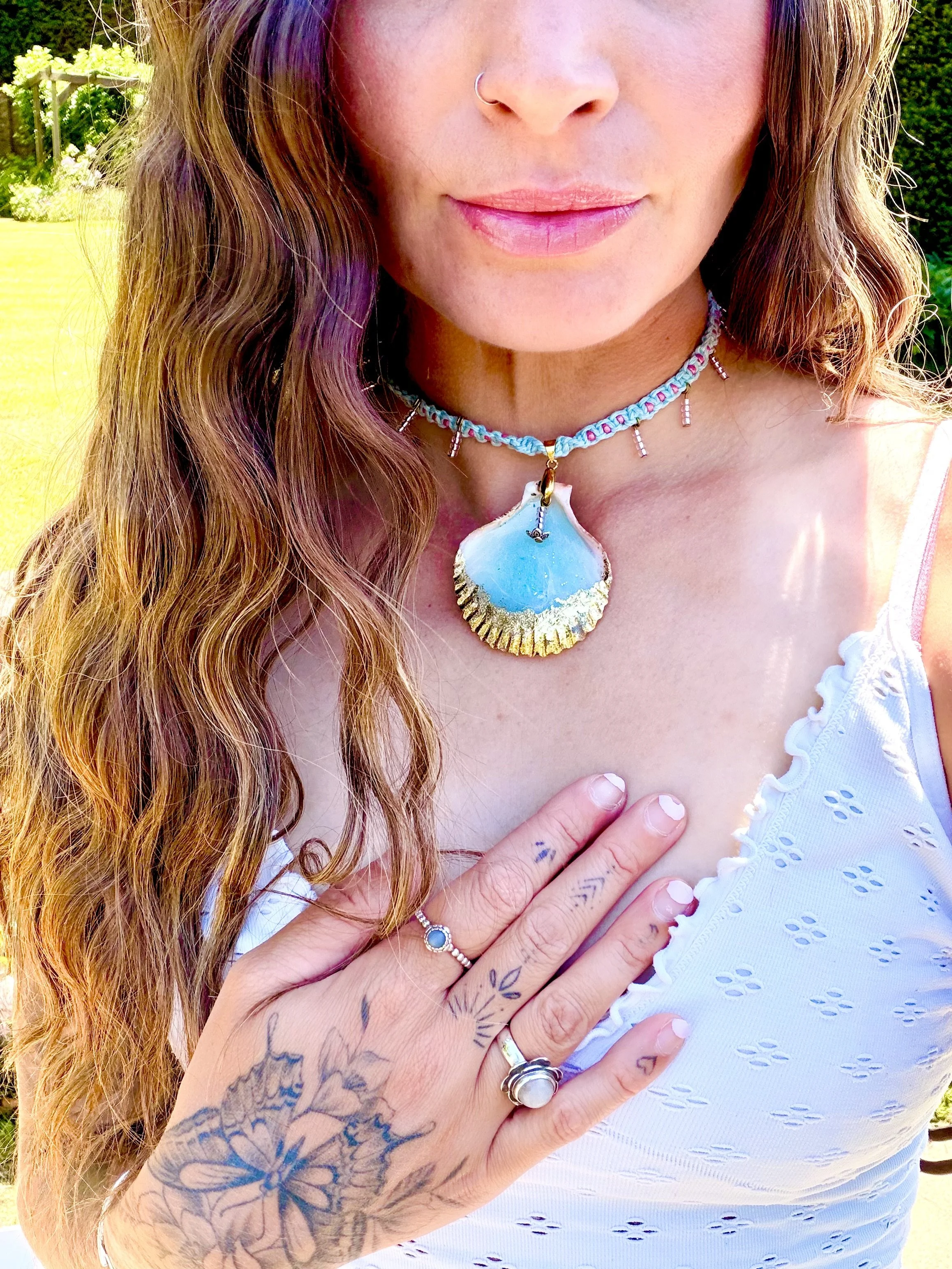 Close-up of a woman's face and upper body showing long wavy hair, necklace with a large blue shell pendant, rings, and tattoos on her hand, with a garden background.